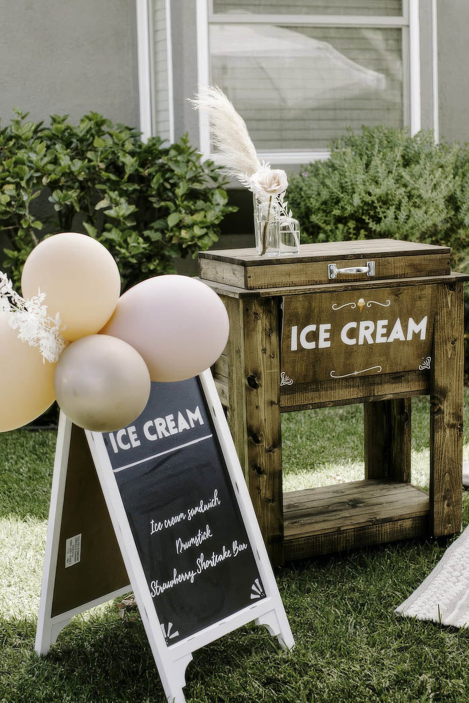 A rustic ice cream stand outdoors, perfect for a boho baby shower, features a wooden chest with "Ice Cream" inscribed on it, a glass jar topped with decorative dried grass, and a chalkboard menu listing ice cream sandwiches, popsicles, and strawberry shortcake bars. Balloons are tied to the stand.