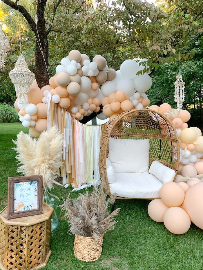 A cozy outdoor setup with a wicker egg chair and white cushions under a tree sets the perfect scene for a Boho baby shower. Surrounding the chair are clusters of beige, white, and peach balloons with hanging macrame decor. Dried pampas grass in vases adds a natural touch. A framed sign is visible on the left.