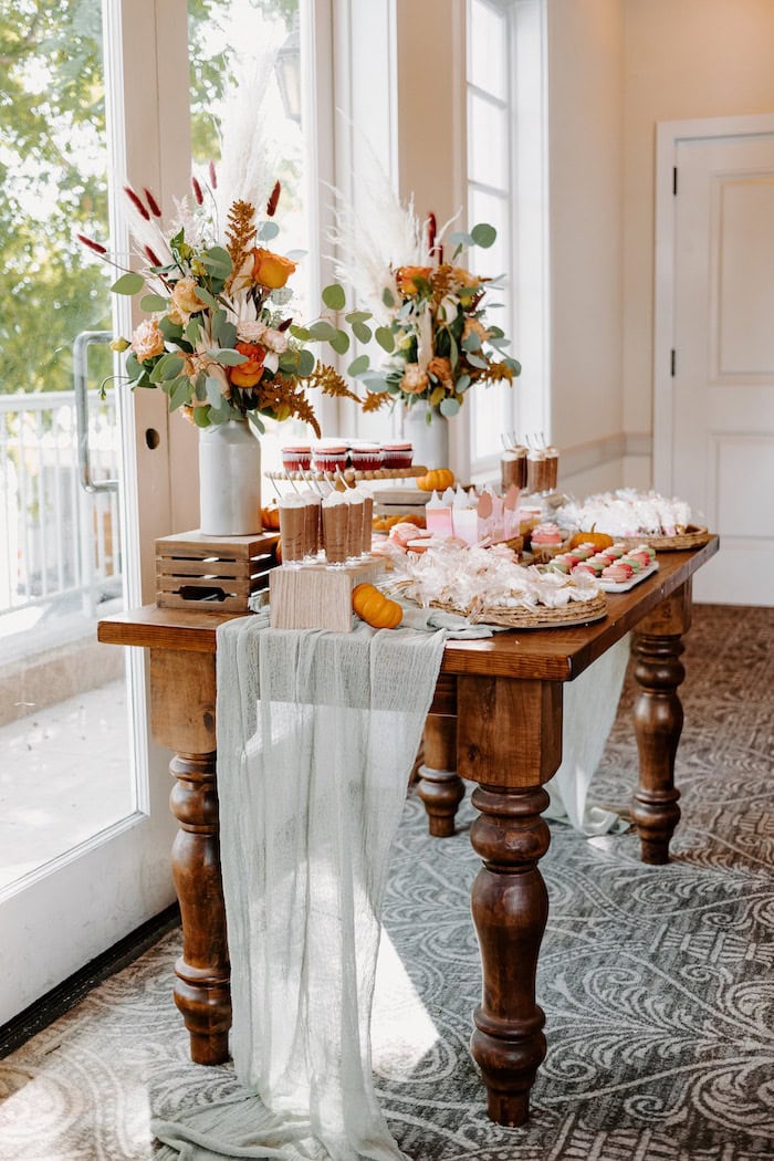 A rustic wooden table set with various desserts, including cakes and pastries, arranged neatly for a boho baby shower. Two vibrant floral arrangements are placed at the back corners of the table, alongside pumpkins and decorative fabric draping over the edge. The background features large windows.
