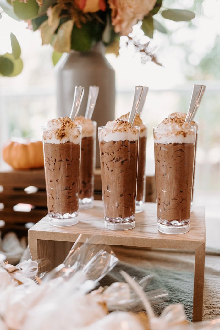 Five tall glasses filled with chocolate mousse topped with whipped cream and sprinkled with cocoa powder. Each glass has a small plastic spoon. The glasses are arranged on a wooden stand, surrounded by flowers and a small pumpkin—a perfect dessert display for a boho baby shower.