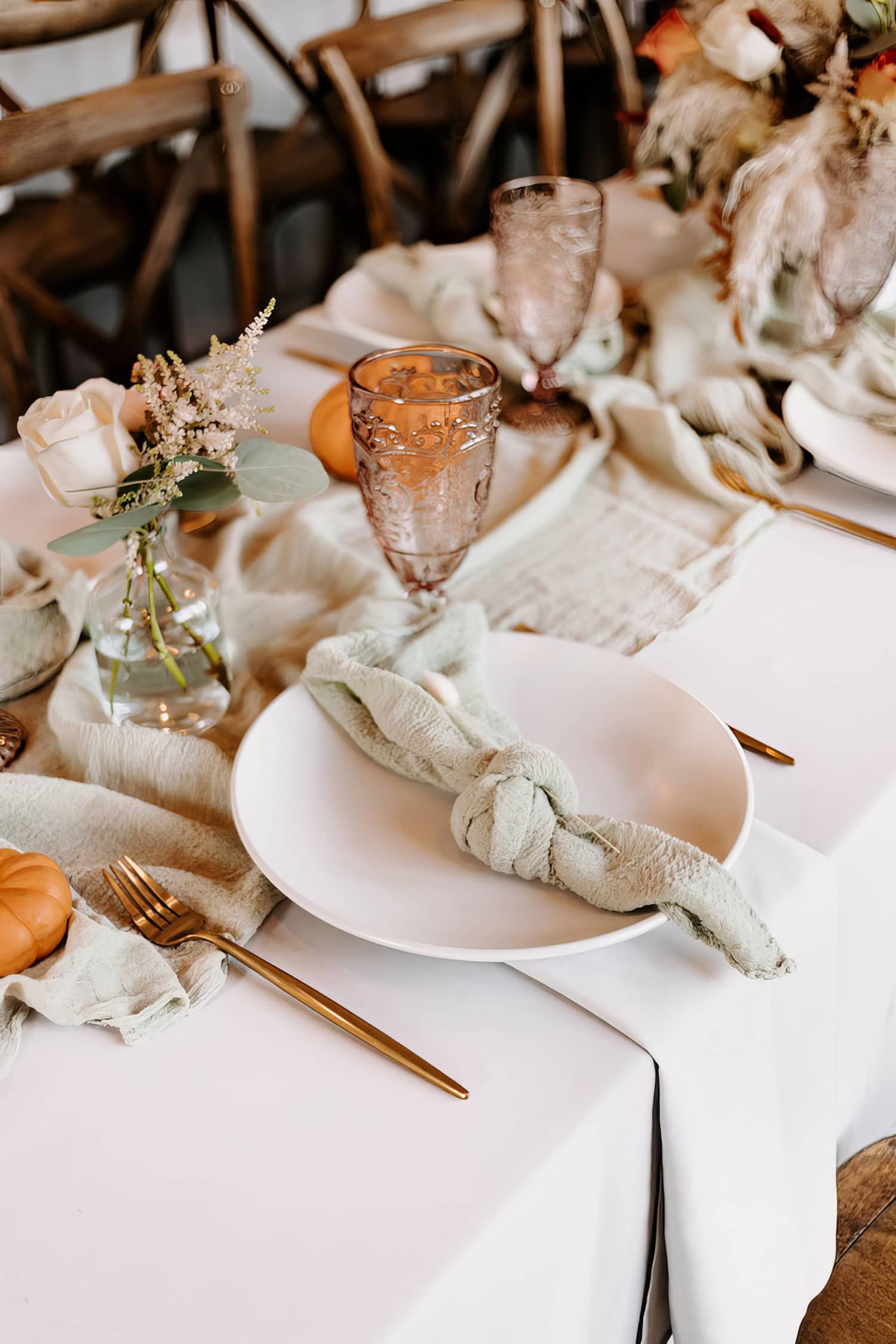 A close-up of an elegantly set table for a Boho baby shower with a white plate, silverware, and a green napkin tied in a knot. Decorative elements like a small vase with flowers, a gourd, and an ornate glass are present. Pale green and white table linens adorn the table.