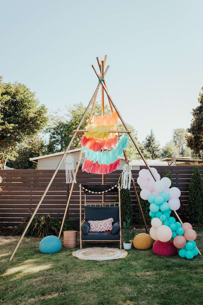 A backyard setup for a Boho baby shower features a wooden teepee adorned with colorful hanging tassels and dreamcatchers. Beneath it, there's a small dark chair, a patterned cushion, and various round floor cushions. To the side, a balloon arrangement includes pastel and bright colors.