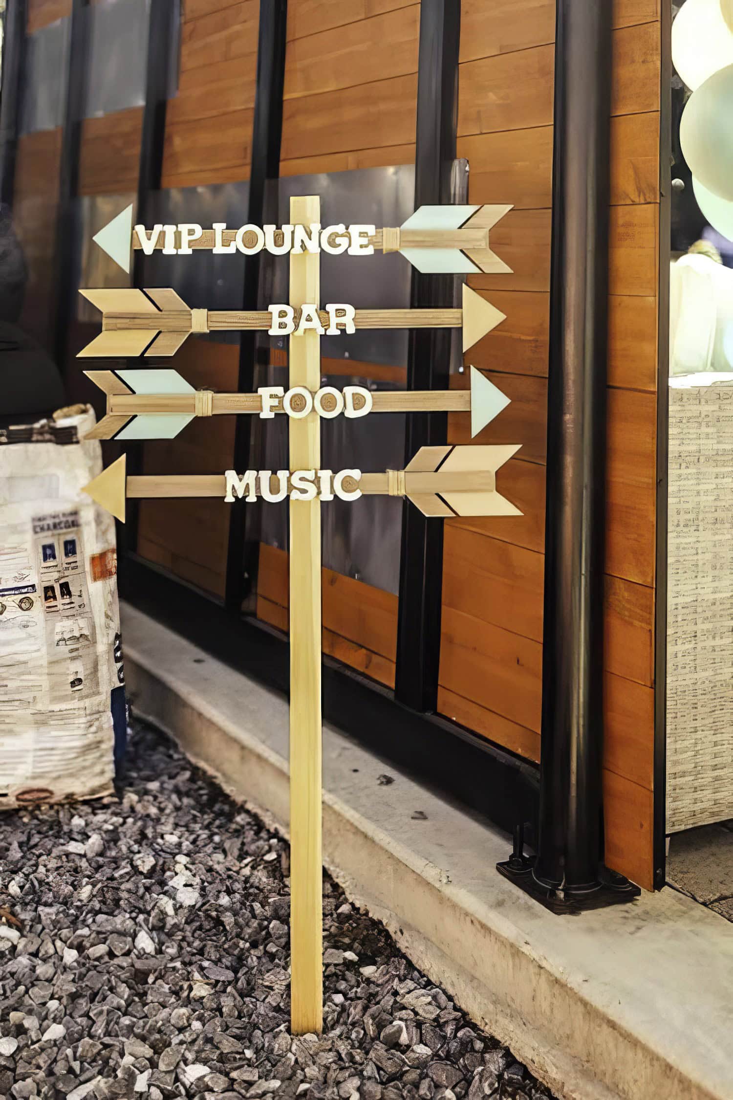 A wooden signpost with arrows pointing in different directions indicating "VIP Lounge," "Bar," "Food," and "Music" stands outside a building. The background features a wooden wall and a small pile of gravel near the base of the signpost, perfect for setting the scene at a Boho baby shower.