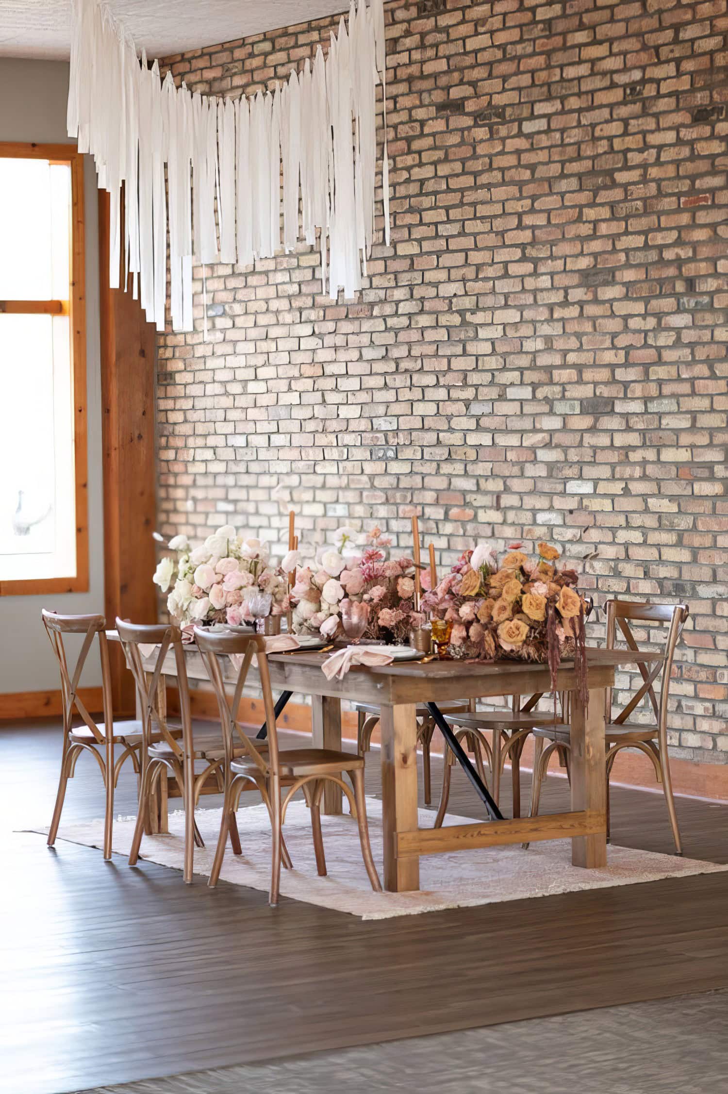 A rustic dining table set with elegant floral arrangements featuring pink, white, and peach flowers, perfect for a Boho baby shower. Six wooden chairs surround the table. The backdrop is a brick wall adorned with white streamers hanging from the ceiling above. Natural light illuminates the scene.