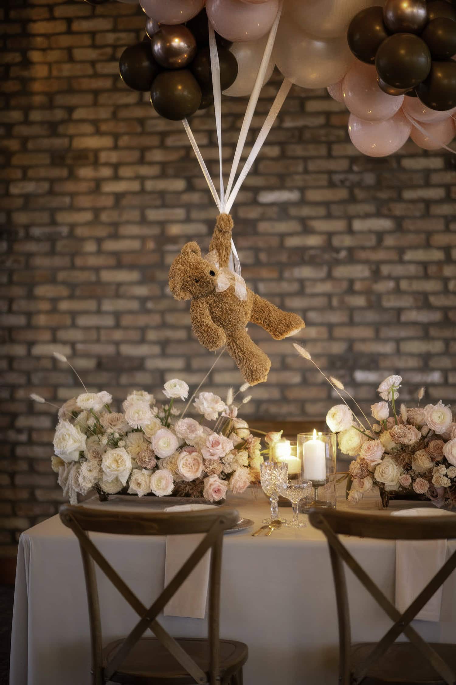 A cozy dining table setup features elegant floral arrangements with white and pink roses, set against a brick wall backdrop. Perfect for a boho baby shower, a teddy bear is suspended above the table, tied to brown, pink, and gold balloons, adding a whimsical touch.