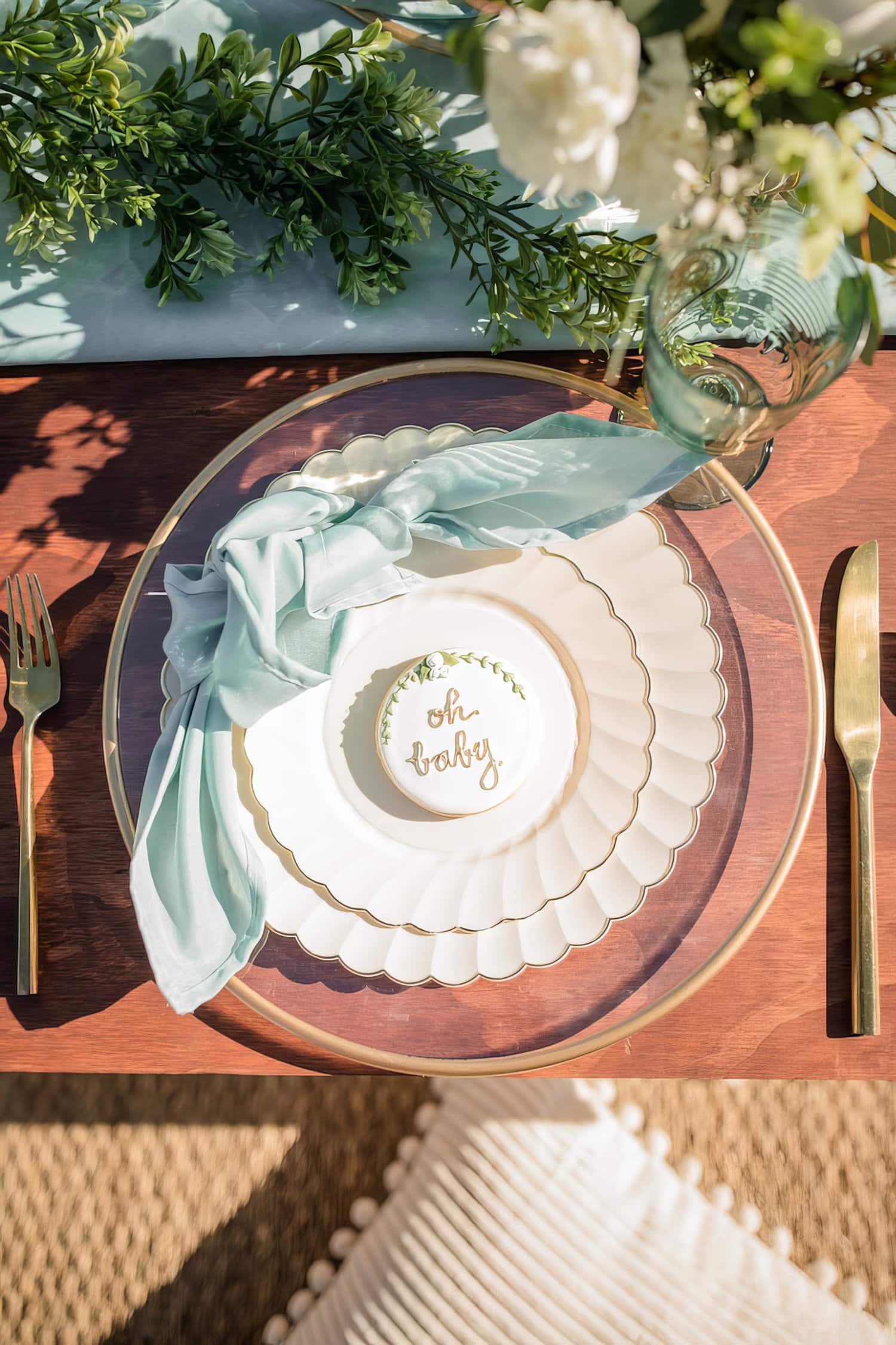 A beautifully set table for a boho baby shower features a white scalloped plate with a smaller dessert plate atop it, showcasing the words "oh baby" in gold. A mint-green napkin drapes elegantly over the plates, and golden cutlery flanks the setting, complemented by lush greenery and flowers.