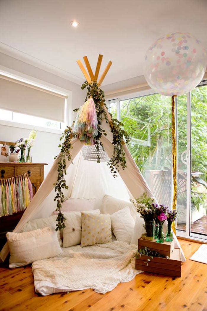 A cozy indoor teepee set up in a bright room with large windows, perfect for a boho baby shower. The teepee is decorated with hanging foliage, a tassel garland, and filled with soft cushions and blankets. A large balloon with confetti inside is tied to one side, and a bouquet of flowers sits nearby.