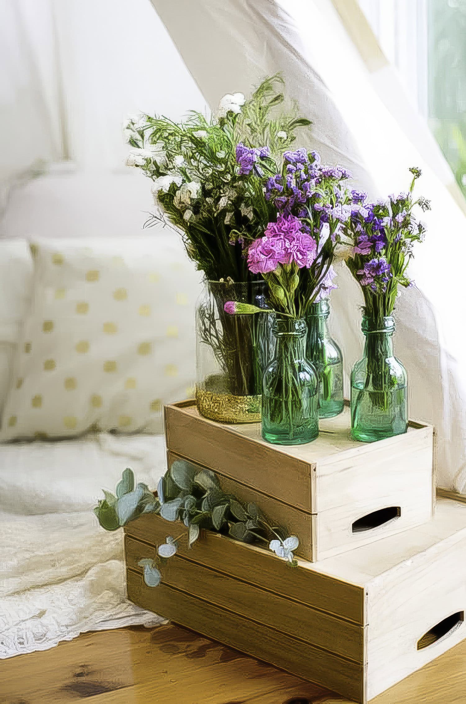 A cozy indoor setup for a boho baby shower featuring two stacked wooden crates displaying three green glass vases filled with vibrant purple and white flowers. Eucalyptus leaves adorn the bottom crate. In the background, a bed with white and yellow polka dot pillows is visible.