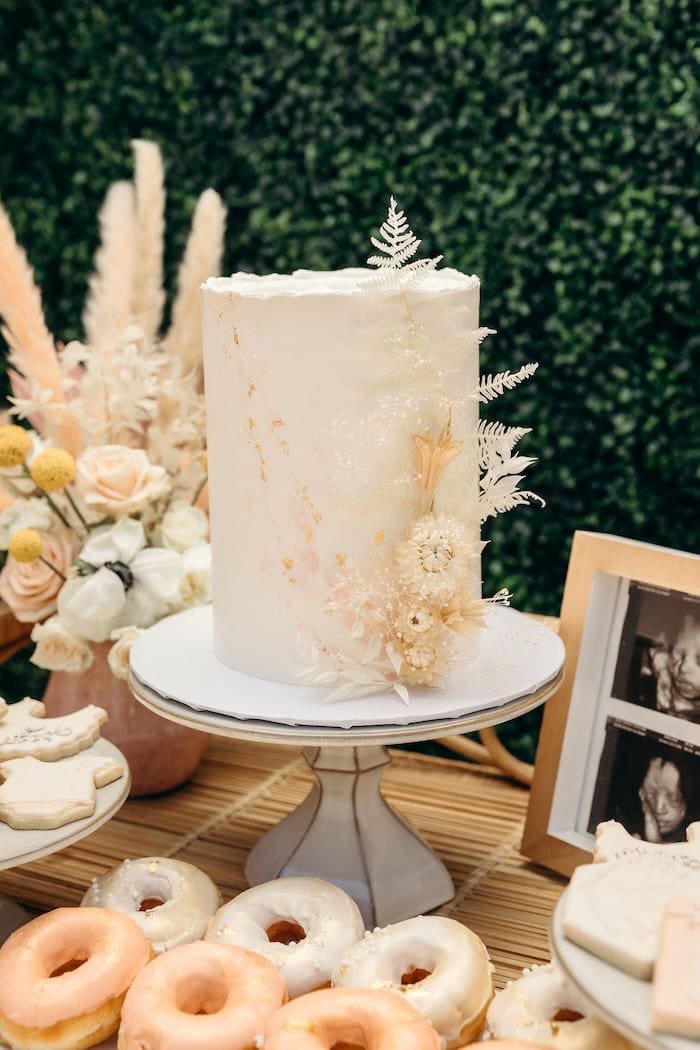 A tall, elegant white cake with subtle floral designs sits on a white cake stand. The cake is adorned with delicate dried flowers and greenery, perfect for a boho baby shower. Surrounding the cake are various doughnuts and cookies decorated with intricate designs, all set on a wooden surface.