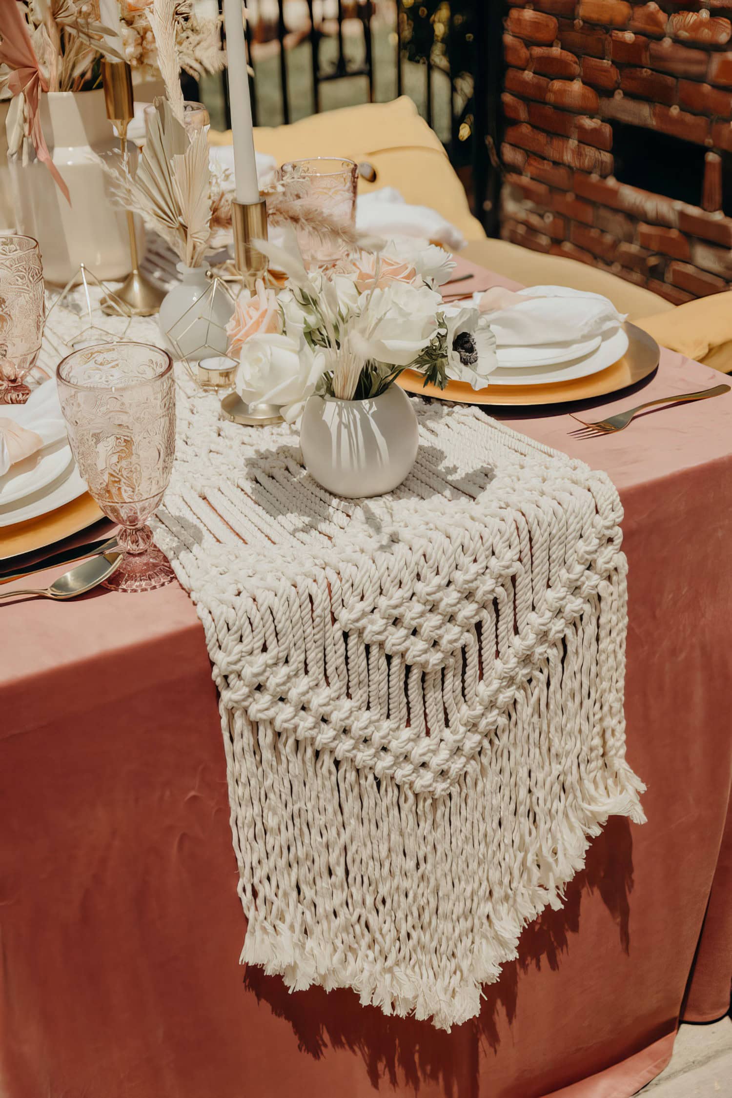 A pink table is elegantly set for a boho baby shower, adorned with white plates, gold-rimmed glasses, and cutlery. A white macramé runner lies down the center, topped with a white vase holding white and peach roses. Tall white candles in gold holders complement the decor.