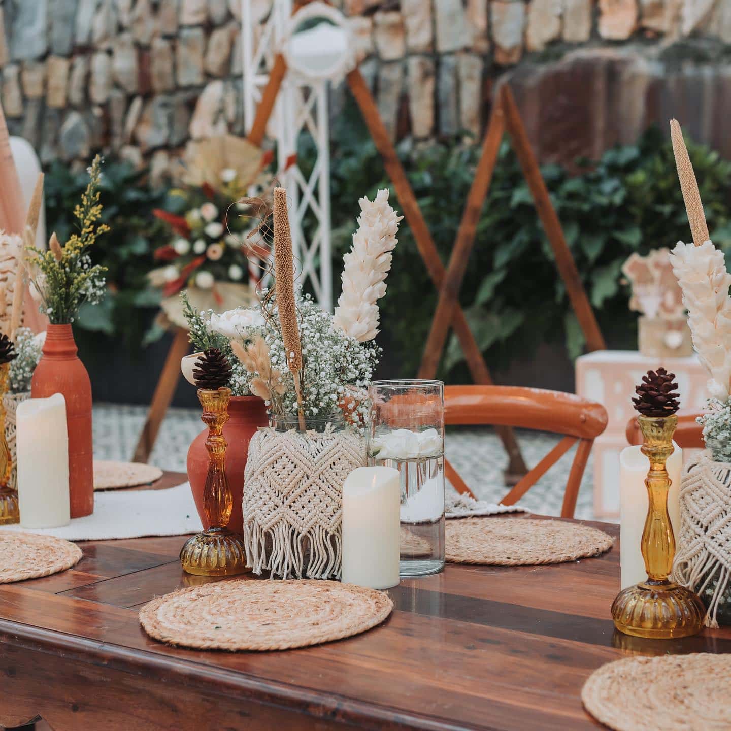 A rustic wedding table setup featuring woven placemats, dried floral centerpieces in ceramic vases, and decorative candles. The background includes a stone wall adorned with greenery and natural wood elements, creating a cozy, elegant ambiance perfect for a boho baby shower.
