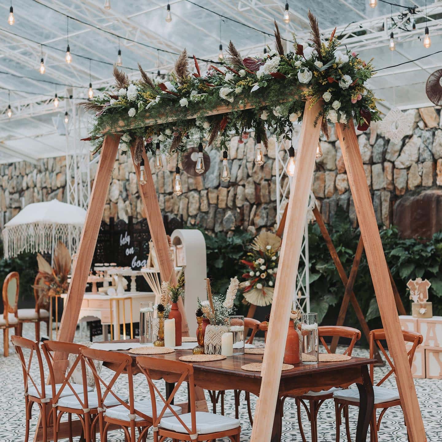 A beautifully decorated outdoor event space perfect for a boho baby shower features a wooden table with chairs and hanging floral arrangements. The setting includes string lights, greenery, and rustic decor elements against a stone wall backdrop, creating a charming, cozy atmosphere.