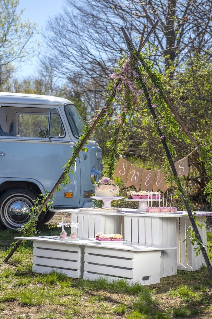 A bohemian-style outdoor dessert table for a boho baby shower features a sweet sign, flowers, and greenery set against the backdrop of a vintage blue van. The table, made of white crates, holds cakes, cupcakes, and other sweets. Trees and grass surround the charming scene.