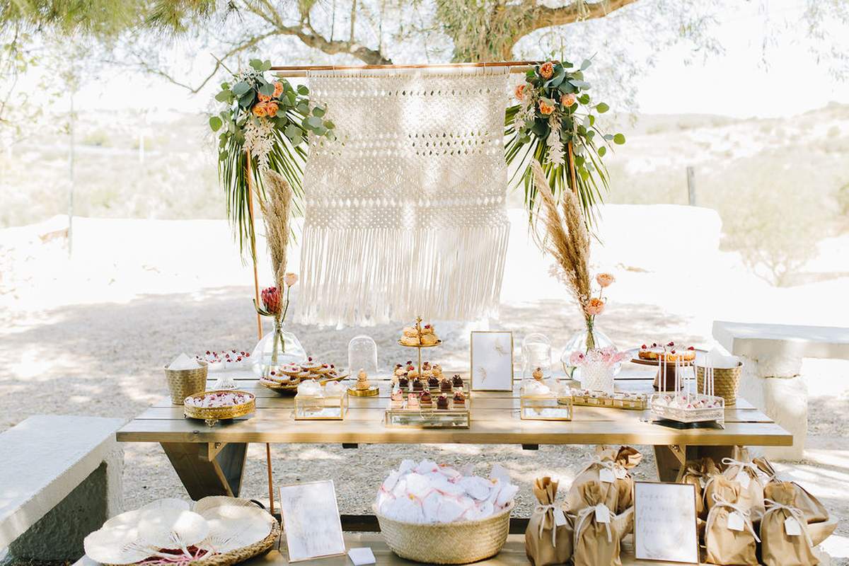 A beautifully decorated dessert table set outdoors for a boho baby shower features a macramé backdrop with floral arrangements. The table holds an assortment of pastries, desserts, and drinks, with party favor bags and large brim hats on display. Trees and a scenic background surround the setup.