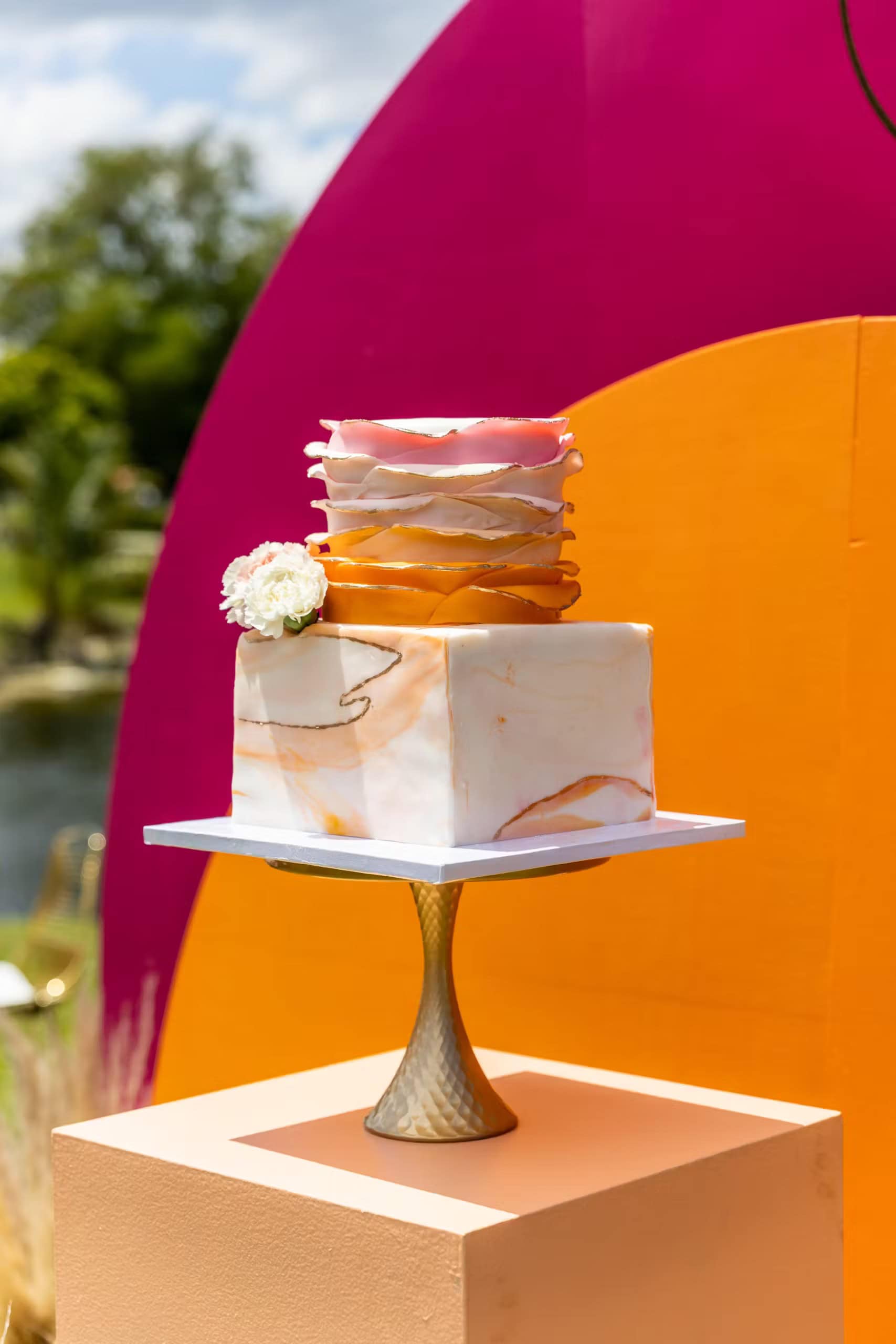 A two-tiered wedding cake stands on a gold pedestal. The bottom tier is a white marble design, while the top tier features pink and orange wavy layers with a white flower embellishment, perfect for a boho baby shower. The background has vibrant pink and orange circular segments with an outdoor setting.