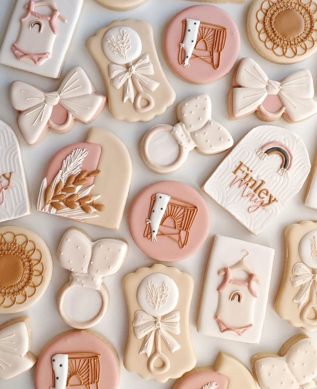 An assortment of decorated cookies in soft beige, pink, and white hues perfect for a boho baby shower. The designs include baby outfits, pacifiers, bows, rainbows, wheat stalks, and floral patterns. The cookies feature intricate detailing and are arranged overlapping on a flat surface.