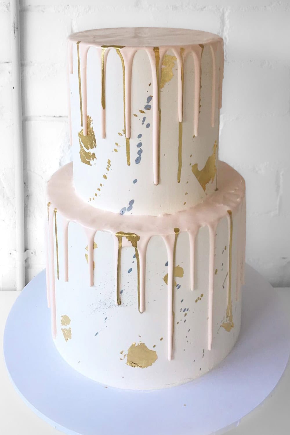 A two-tiered white cake with light pink drip icing flows over the edges of both tiers. Perfect for a boho baby shower, the cake is adorned with gold and silver leaf accents scattered artistically on the smooth, white surface. The cake sits on a white round cake board.

