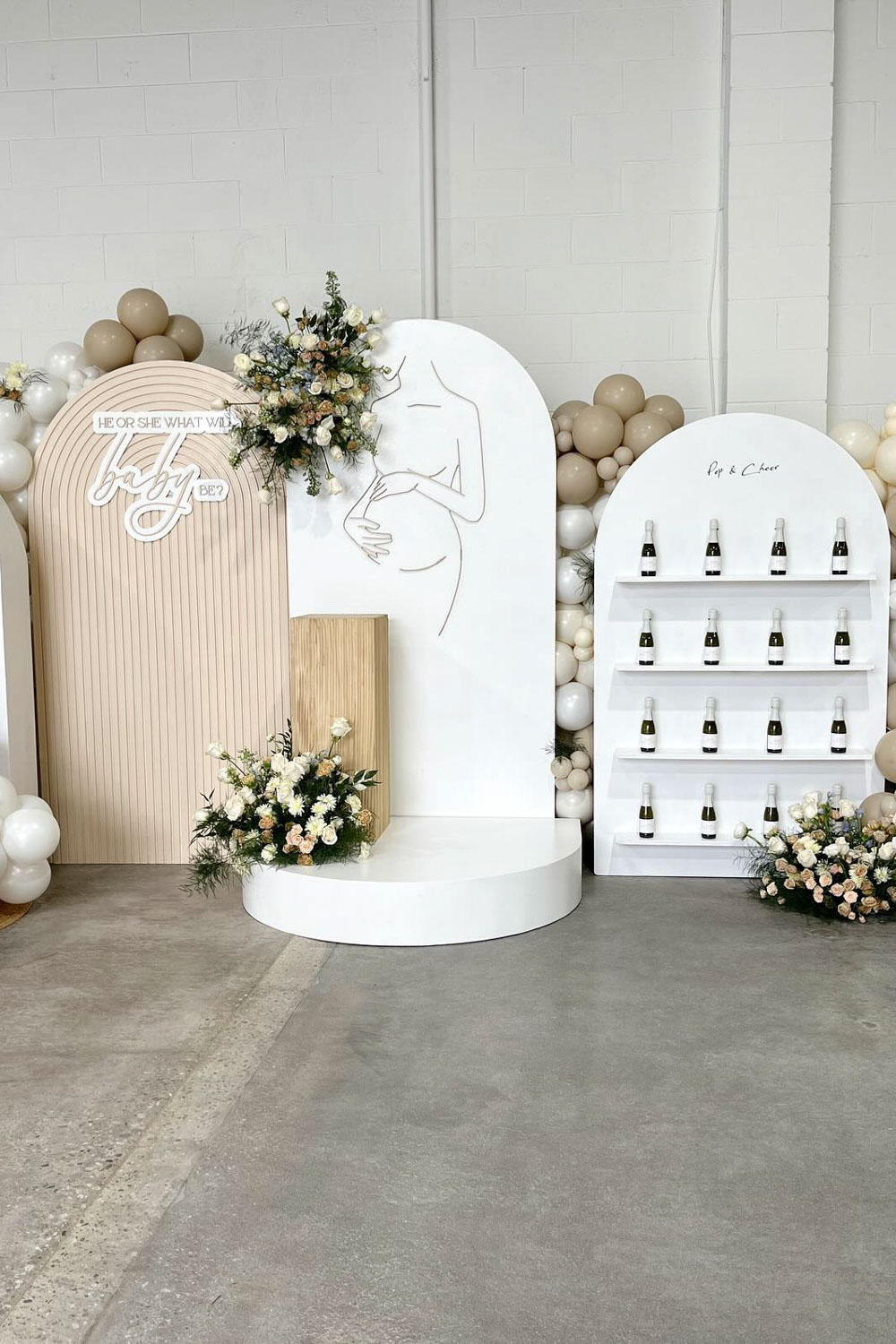 A boho baby shower setup featuring a white-themed decorative backdrop with a floral arrangement and a minimalist line drawing of a pregnant person. Next to it is a display shelf with small bottles and another floral arrangement. Balloons in neutral tones decorate the serene scene.
