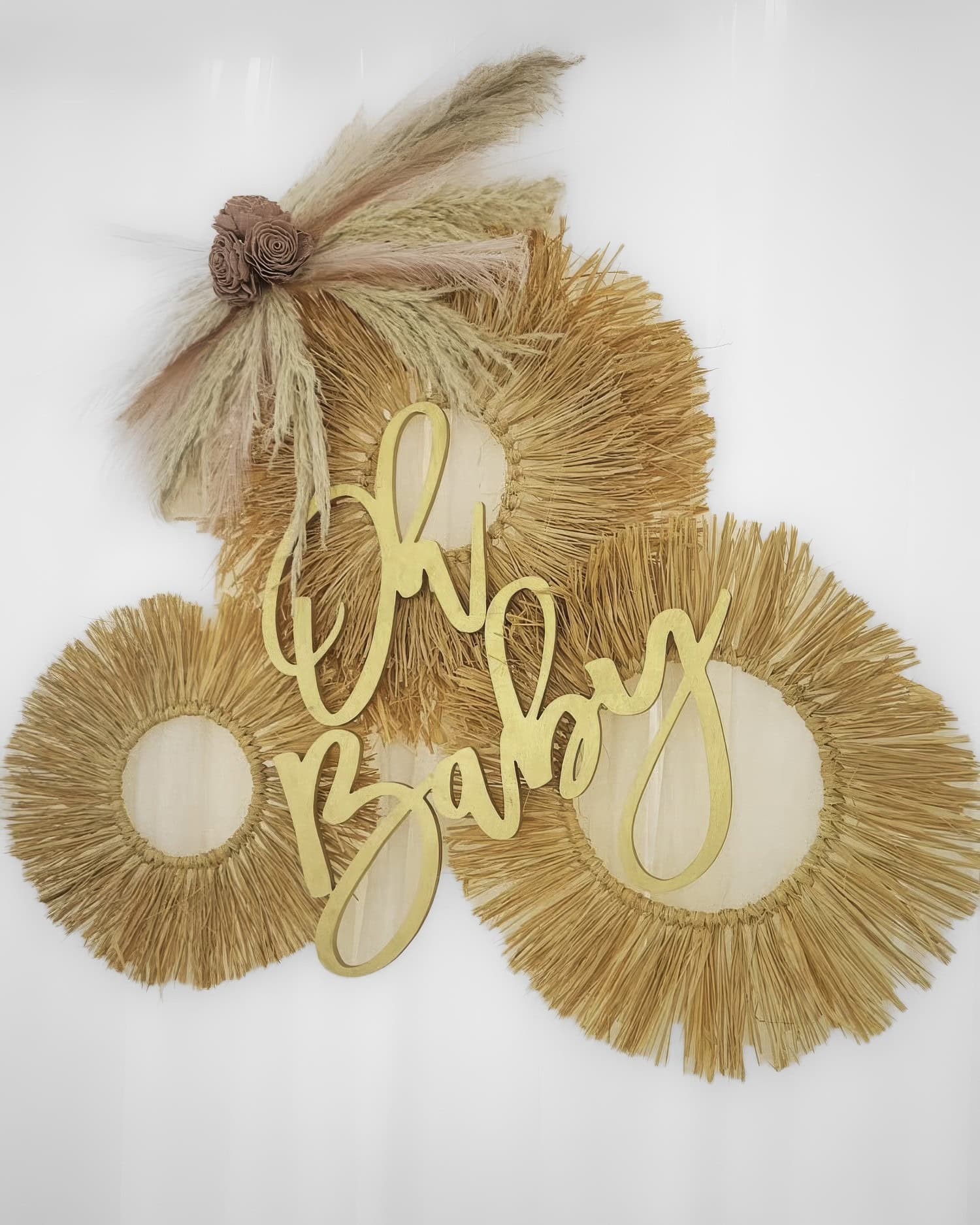 A decorative wall hanging perfect for a boho baby shower features three circular, woven straw wreaths adorned with dried grasses and flowers. In front, a wooden cutout reads "Oh Baby" in cursive script. The neutral tones provide a rustic, bohemian aesthetic.
