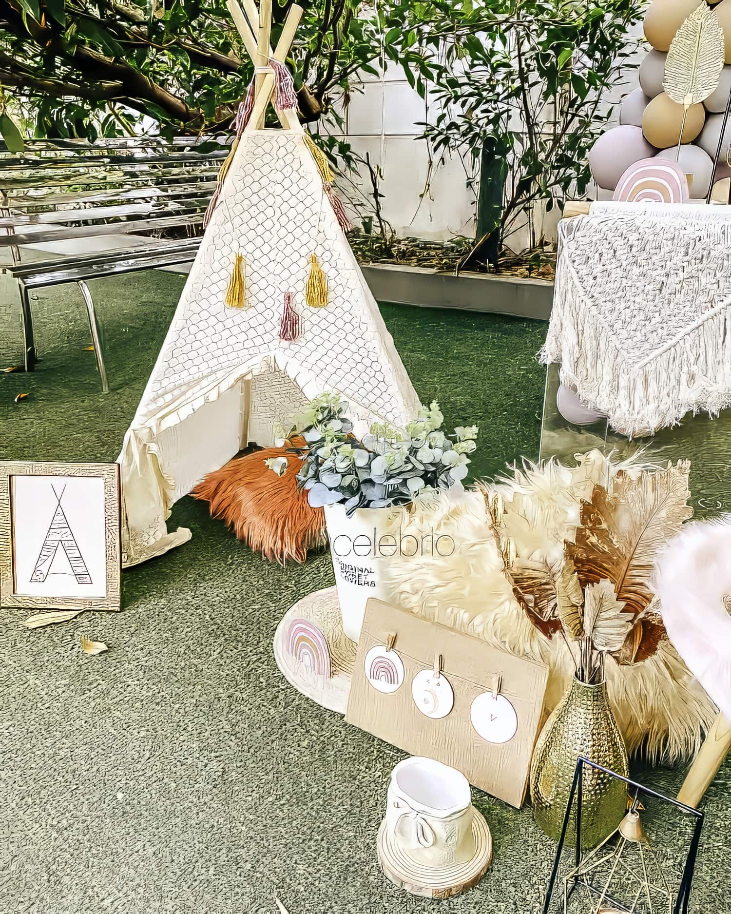 A cozy outdoor setup featuring a small white teepee with a crochet pattern, perfect for a boho baby shower. Decorated with fringe and soft pillows inside, it stands amidst dried plants, feathers, and a "celebrio" sign. Colorful balloons are tied to a table draped with a macrame cloth, completing the serene ambiance.