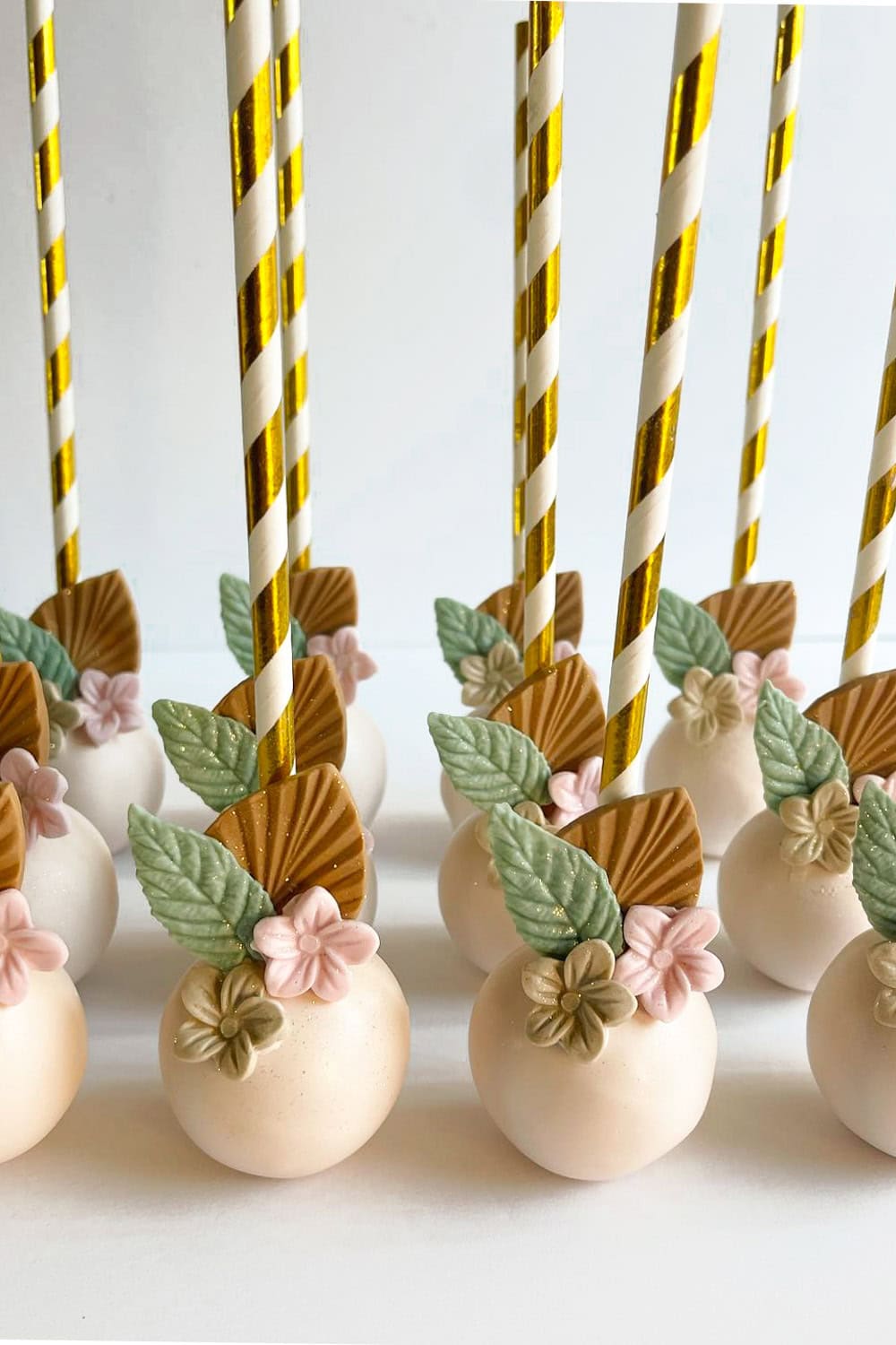 A row of elegant cake pops decorated with pastel pink and beige flowers, green leaves, and golden accents, perfect for a boho baby shower. Each cake pop is held by a white and gold-striped stick, arranged neatly on a white surface.