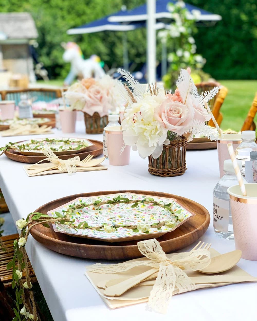 A beautifully set outdoor table for a boho baby shower, featuring wooden chargers, floral-patterned plates, and neatly wrapped bundles of wooden cutlery with lace. Centerpieces of light pink roses and white flowers in wicker baskets adorn the table, set beside bottles of water and paper cups.