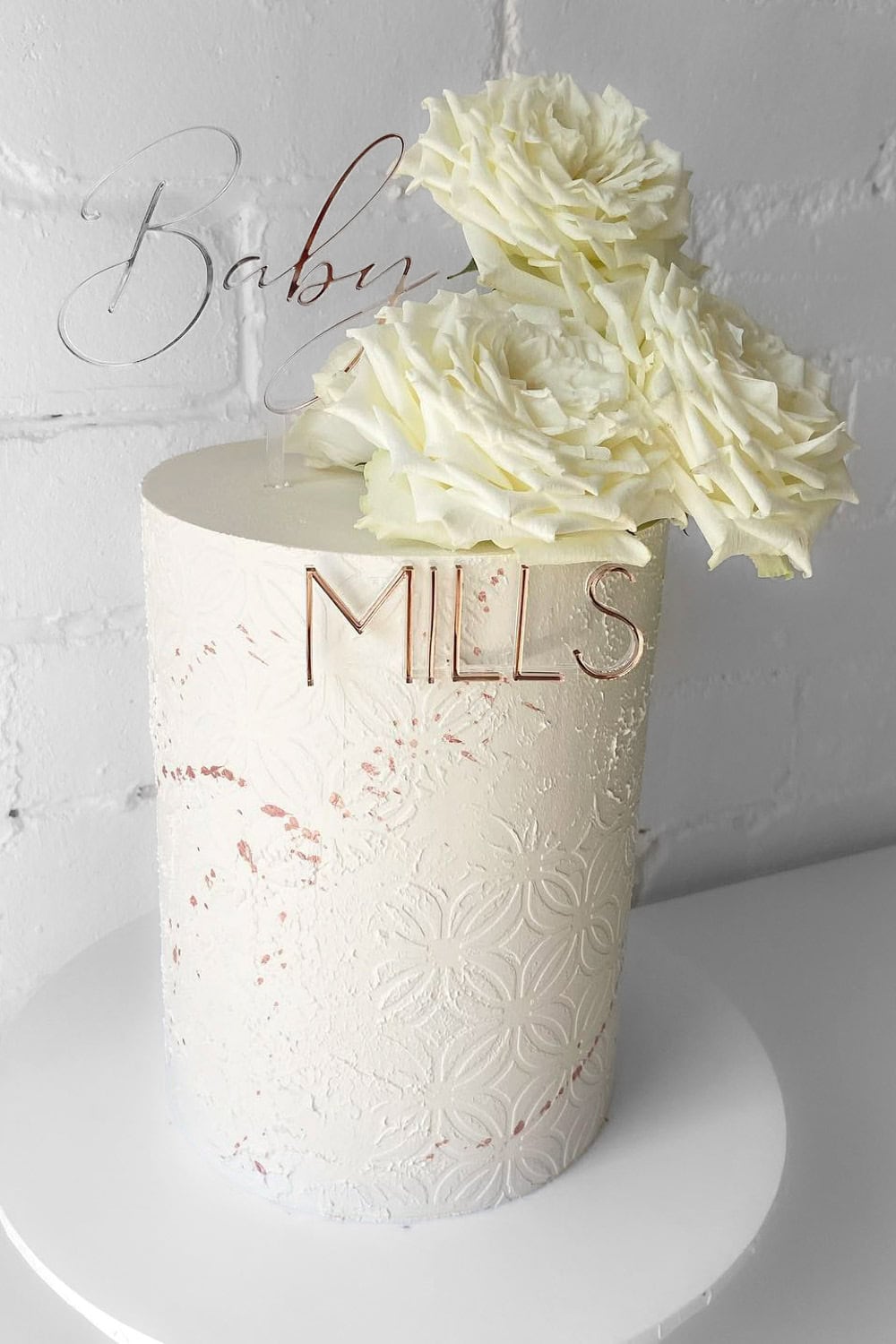 A white baby shower cake with intricate floral patterns and large white roses on top, perfect for a boho baby shower. The cake features the word "Baby" in a clear, elegant script on top and "MILLS" in metallic font on the side. It sits gracefully on a simple white round base.