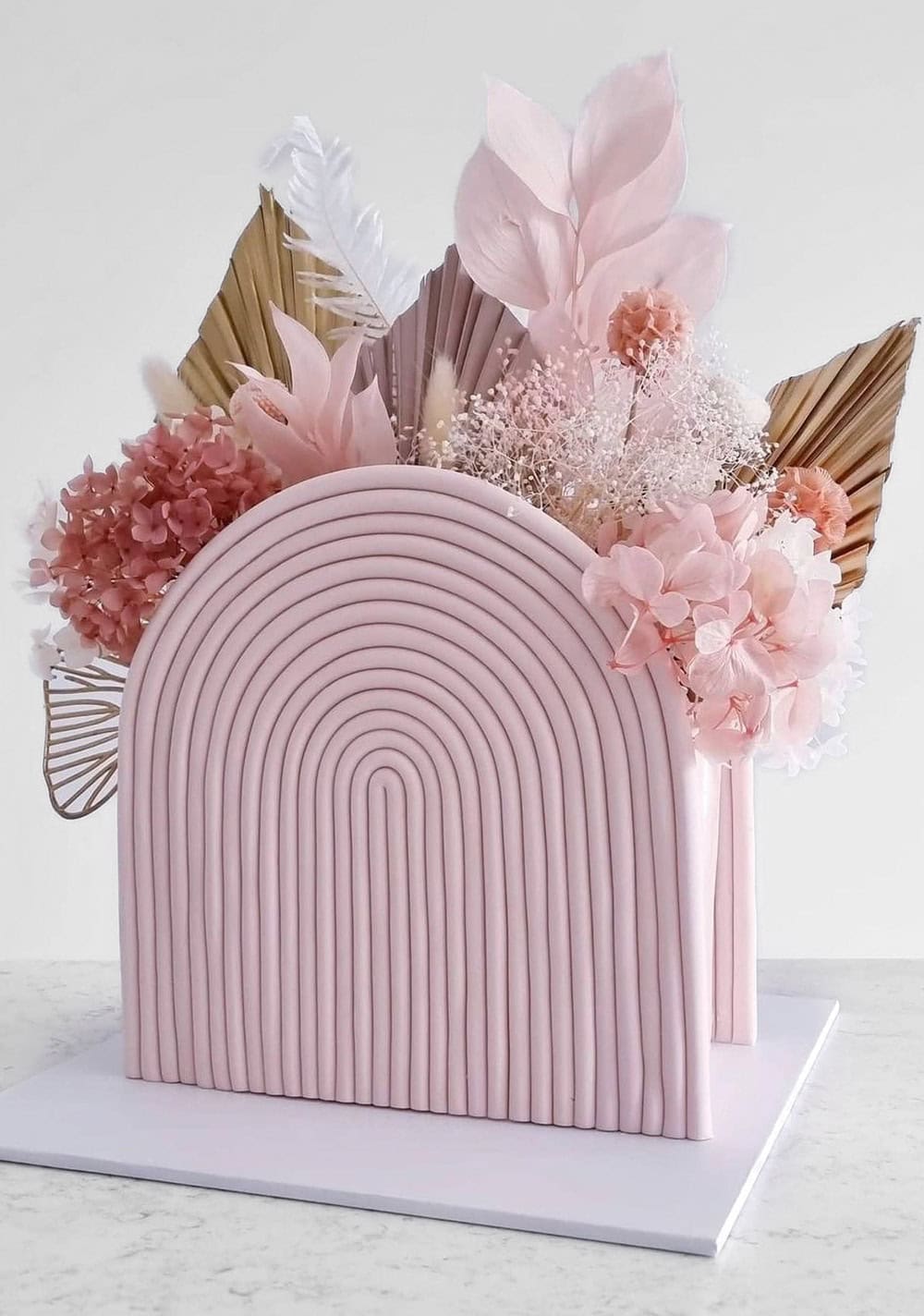 A minimalist pink cake with a unique arched top design, perfect for a boho baby shower. The cake is adorned with dried botanical elements, including pale pink flowers, leaves, and pampas grass, giving it an elegant and contemporary look. It sits gracefully on a white base.