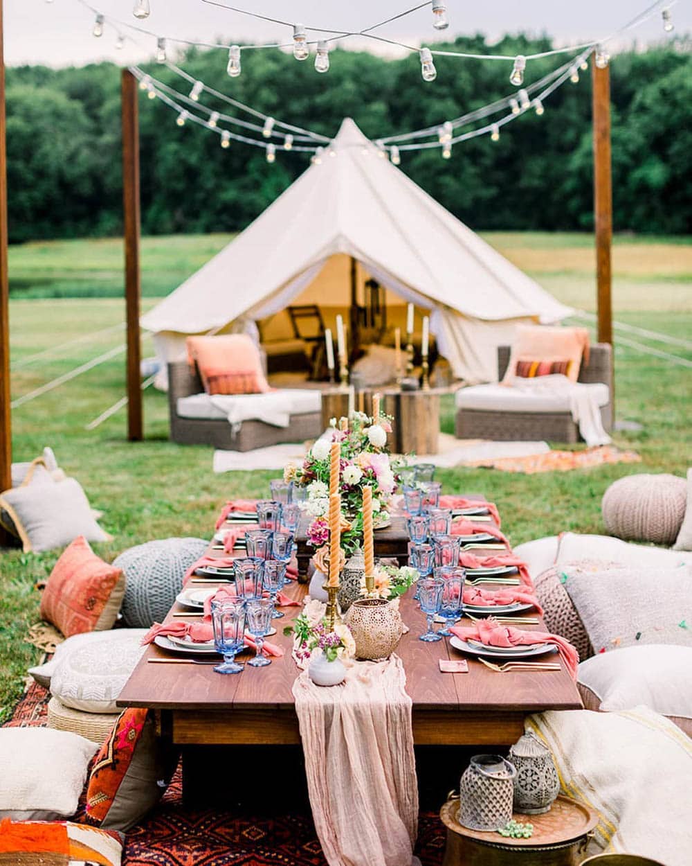 A beautifully decorated outdoor picnic setup perfect for a boho baby shower, featuring a low wooden table adorned with pink table runners, candles, flowers, and blue glassware. Cushions and poufs surround the table, with a cozy tent and more seating in the background. Twinkling string lights hang above, set against lush greenery for a picturesque backdrop.