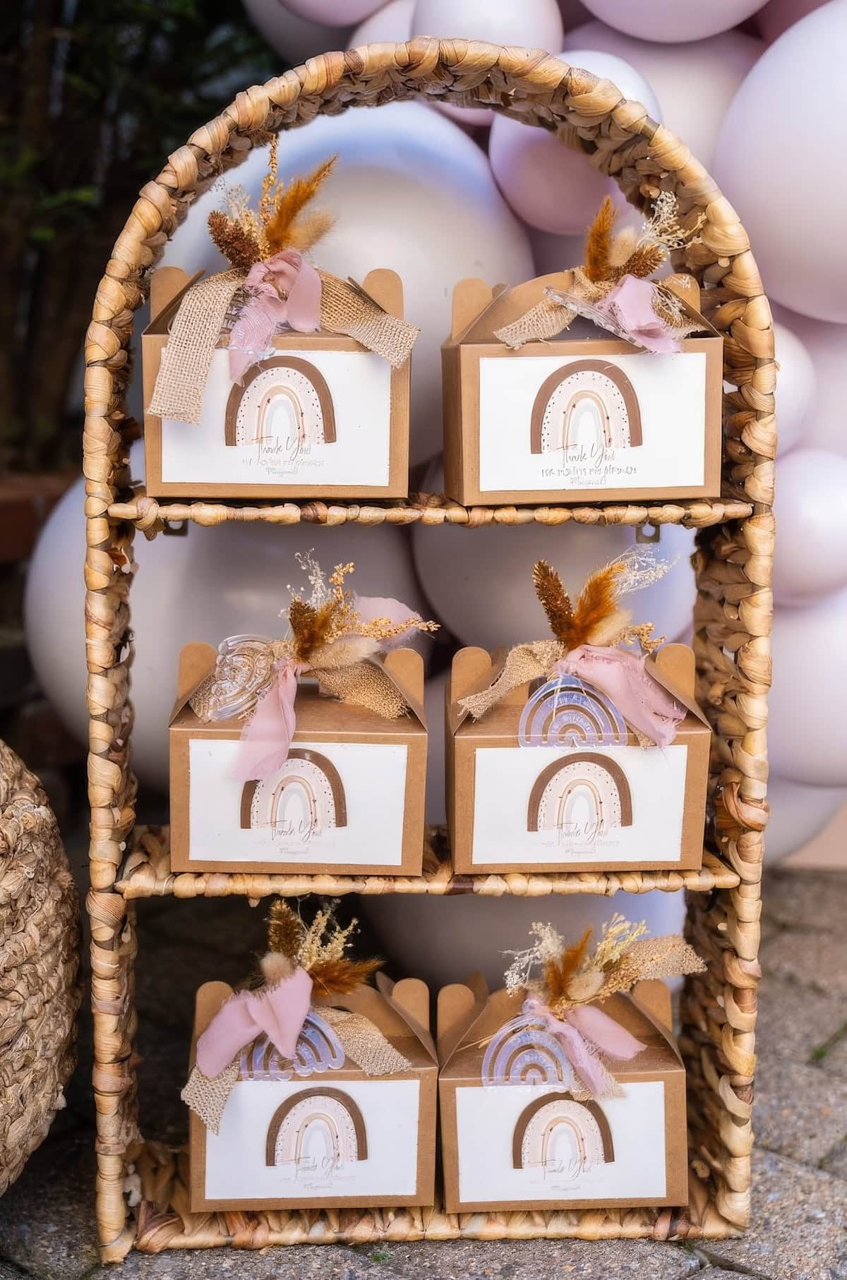 A woven wicker shelf holds six gift boxes adorned with dried flowers and soft pink ribbons, perfect for a boho baby shower. Each box features a label with a rainbow design. The background includes a cluster of white and pink balloons, adding to the festive atmosphere.
