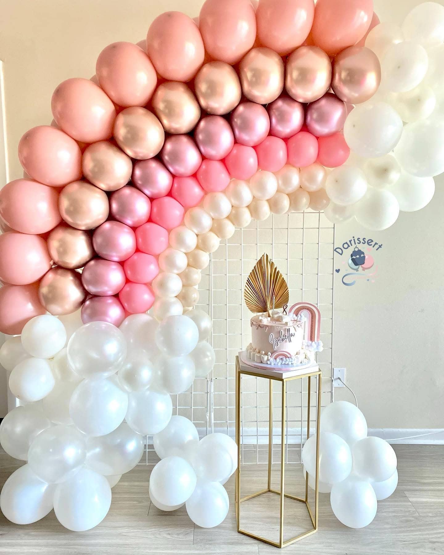 A decorative setup for a boho baby shower featuring a rainbow balloon arch arranged in a gradient from dark pink to white. Below the arch is a gold and white cake stand holding a white cake with pink accents and a gold leaf decoration. The background includes a white grid and a logo on the wall.