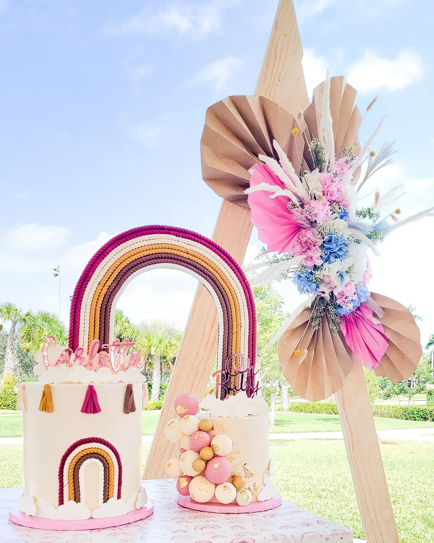 Two white cakes are decorated with a rainbow theme perfect for a boho baby shower. The left cake features a rainbow design and tassels on the side, while the right cake is adorned with pink, white, and gold spheres, and pink toppers that read "Cake" and "Baby." A colorful flower arrangement is displayed nearby.