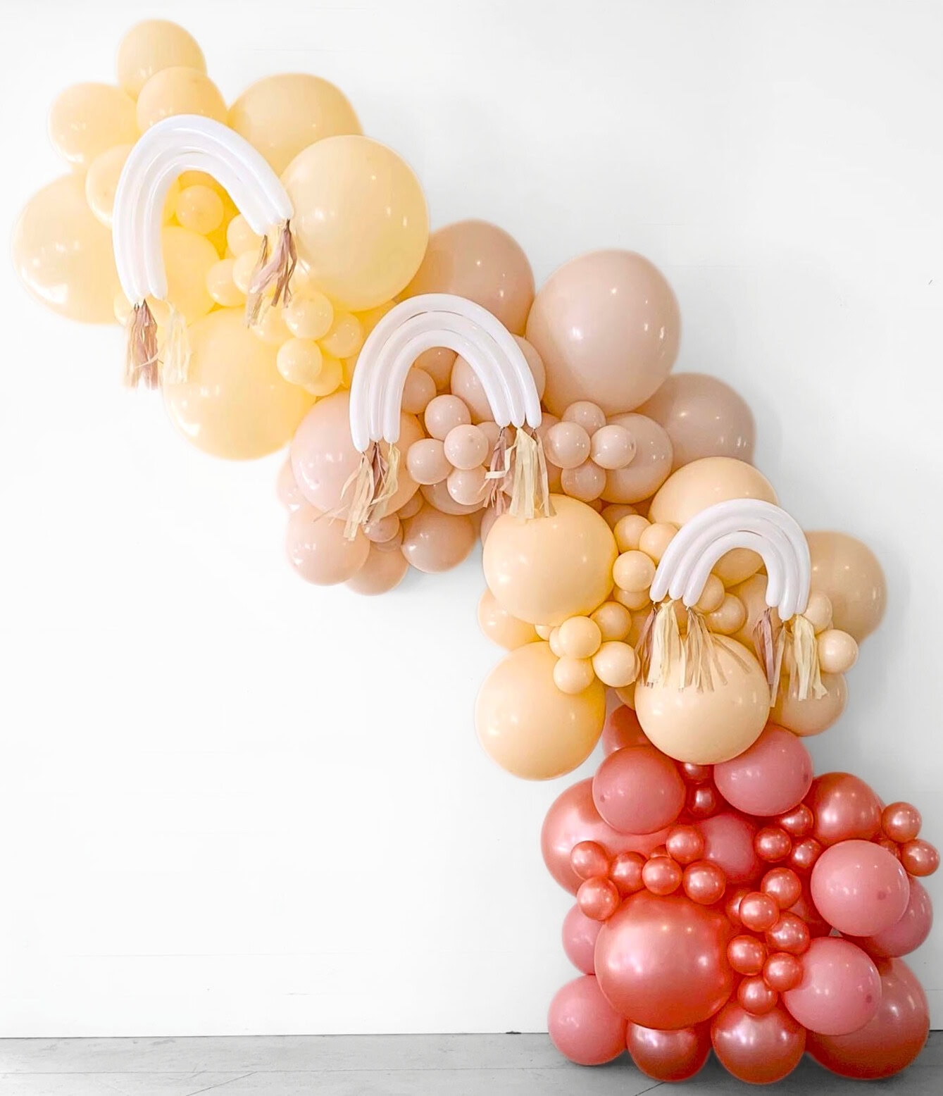 A balloon display perfect for a boho baby shower, featuring a rainbow cascade of balloons in shades of beige, peach, and red. White balloon rainbows with beige tassels are interspersed throughout the arrangement, creating a whimsical and festive decoration against a plain white background.