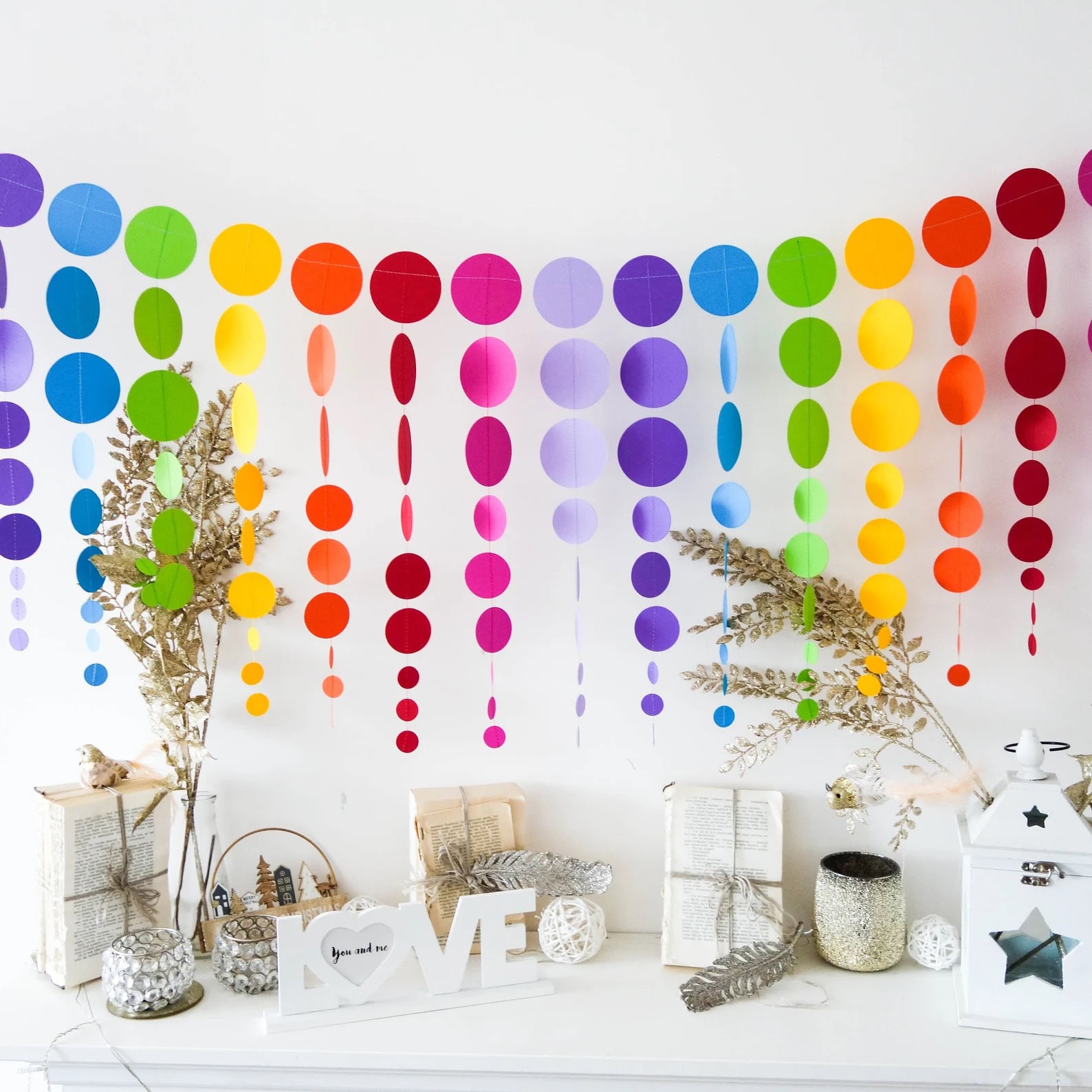 A white shelf decorated with books, dried flowers, lanterns, and a "LOVE" sign is the perfect centerpiece for a rainbow baby shower. Above the shelf hangs a colorful garland made of circles in various sizes and colors, including red, orange, yellow, green, blue, and purple, creating a festive atmosphere.