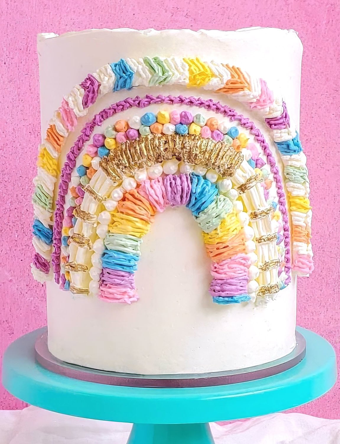 A tall, white frosted cake is decorated with a colorful, textured rainbow design made of buttercream and various icing techniques. Perfect for a rainbow baby shower, the cake features shades of pink, purple, blue, green, yellow, and gold accents and is set on a turquoise cake stand against a pink background.