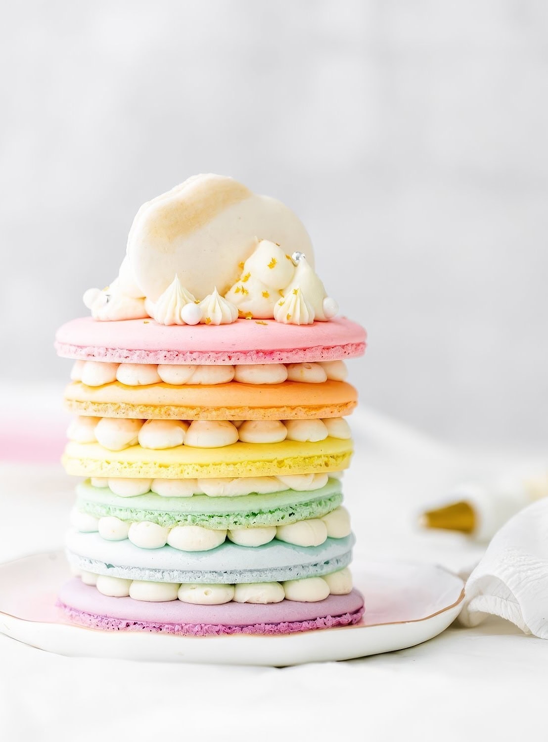 A colorful, layered dessert perfect for a rainbow baby shower, made of pastel-hued macarons in pink, yellow, green, and blue. Dollops of cream sit between each layer. The top is adorned with white frosting and small golden decorations. It sits elegantly on a white plate.