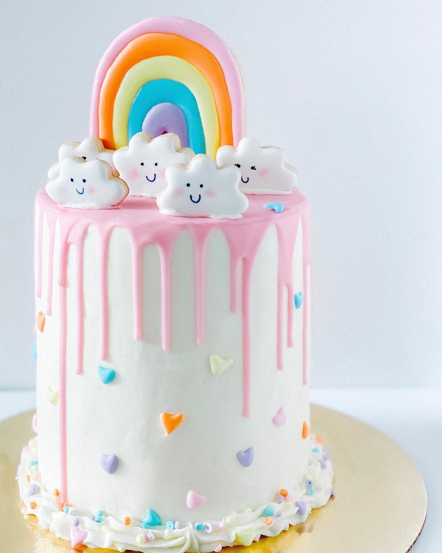 A tall, white cake with pink icing drips down the sides and is topped with a colorful rainbow and smiling cloud decorations, perfect for a rainbow baby shower. The cake is adorned with small, colorful heart-shaped sprinkles and sits on a gold cake board.