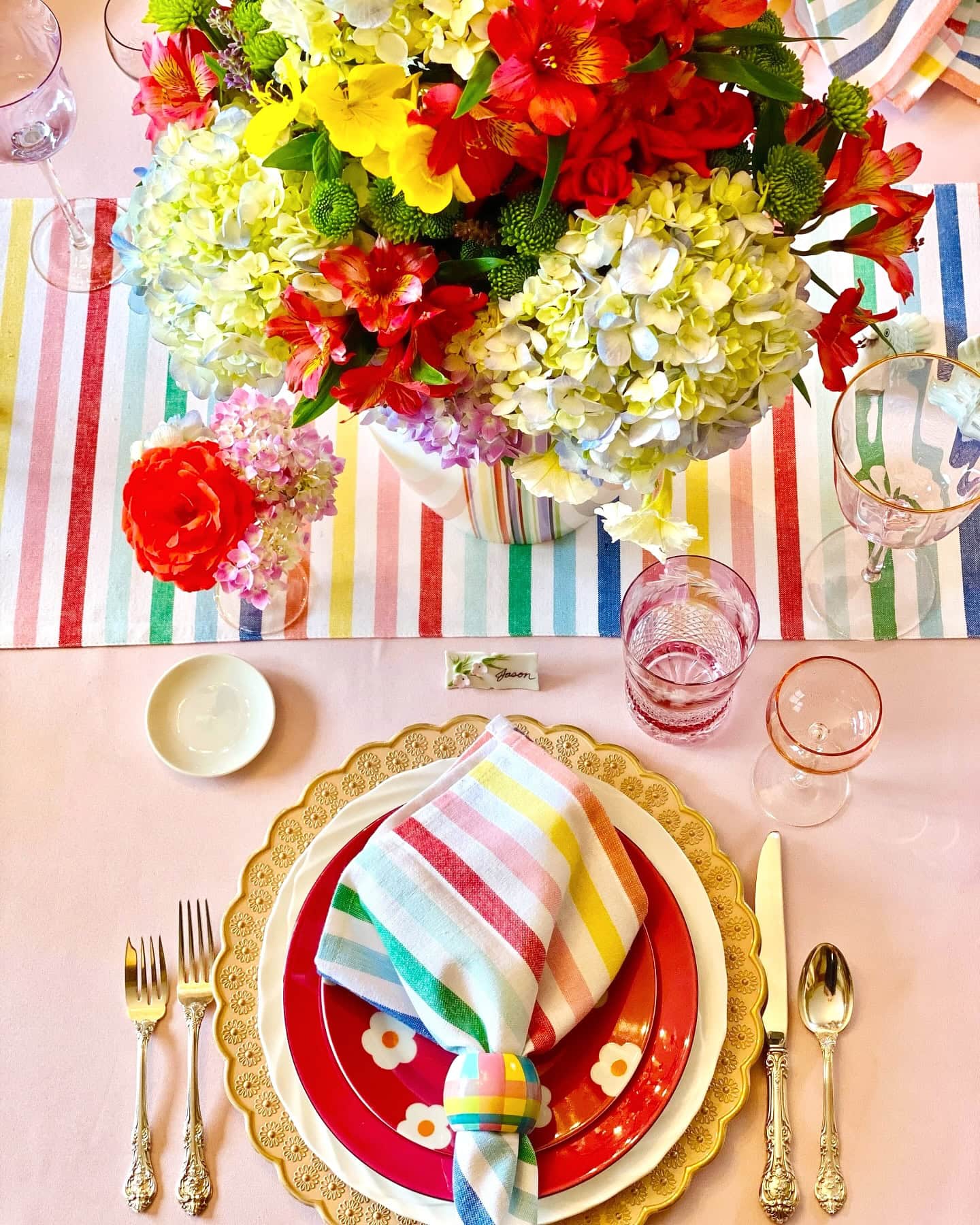 A colorful table setting perfect for a rainbow baby shower features a vibrant floral centerpiece with red, yellow, and white flowers. Gold-rimmed plates with floral designs, rainbow-striped napkins, and pink glassware adorn the table, set on a pastel striped table runner.