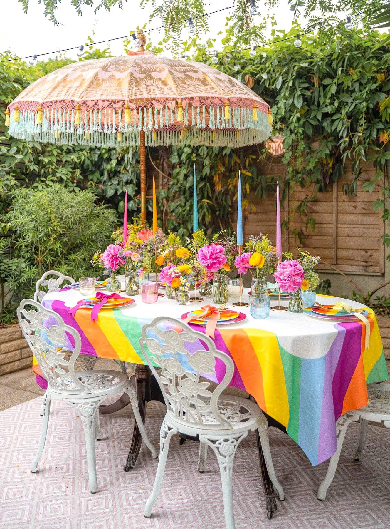 A vibrant outdoor table setting perfect for a rainbow baby shower features a rainbow-striped tablecloth, pink and yellow flowers in glass vases, colorful candles, and place settings with white ornate chairs. An intricate fringed parasol shades the table, surrounded by lush greenery.