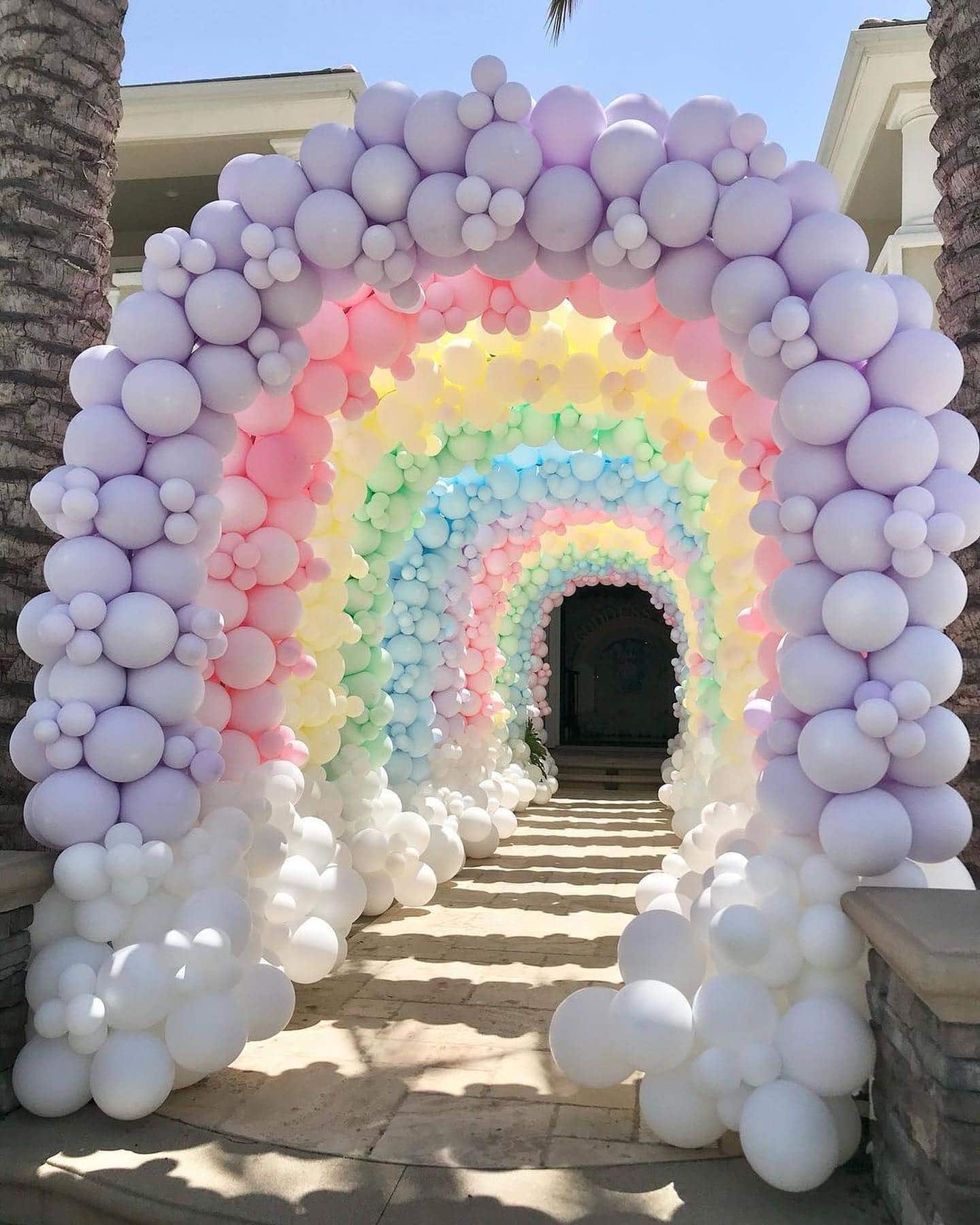 A decorative archway made of colorful pastel balloons, including shades of pink, purple, blue, and yellow, is placed over a pathway leading to a building entrance. Perfect for a rainbow baby shower, the balloons are arranged in a gradient, creating a festive and whimsical rainbow effect.