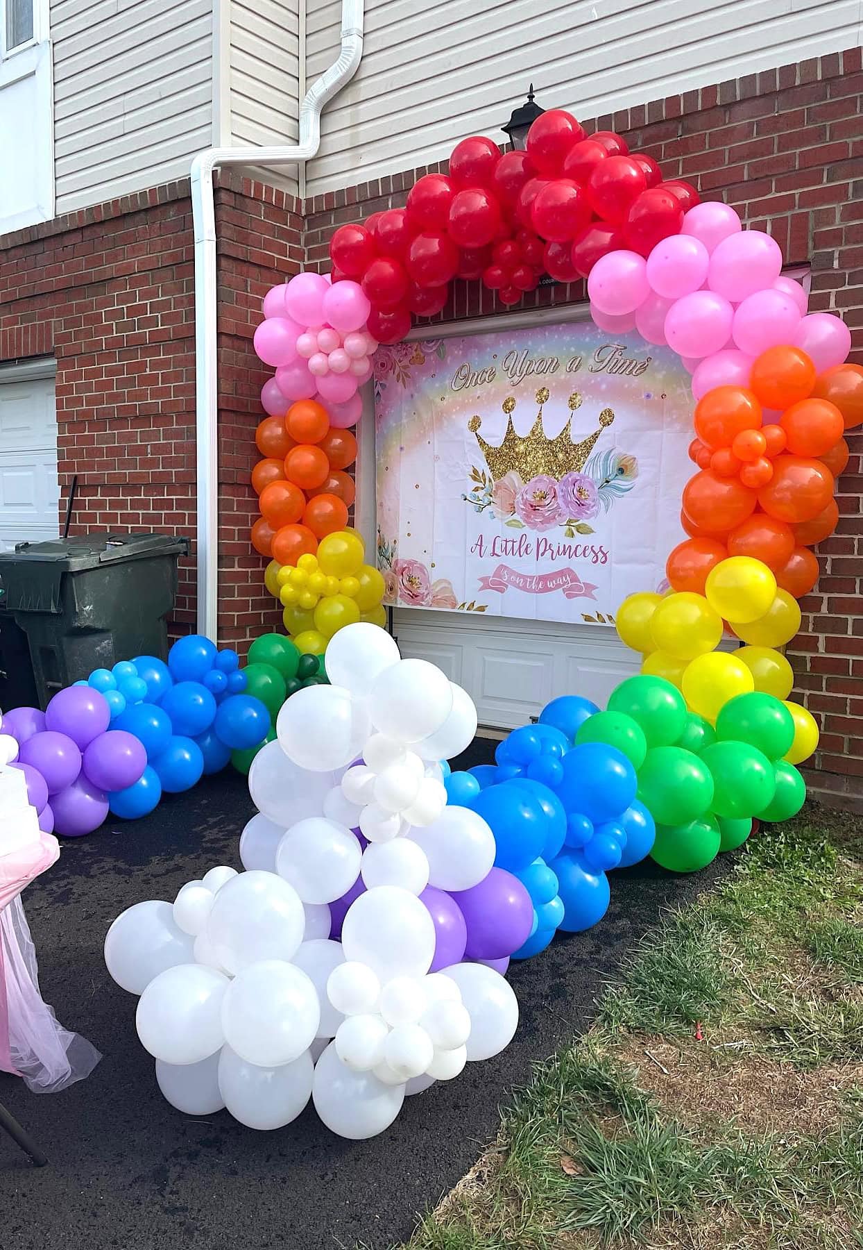 A colorful balloon arch, ranging from red at the top to purple at the bottom, decorates the entrance of a building. Underneath is a banner with a golden crown and the text "Once Upon a Time, A Little Princess." White balloon clusters are placed in front, perfect for a rainbow baby shower celebration.