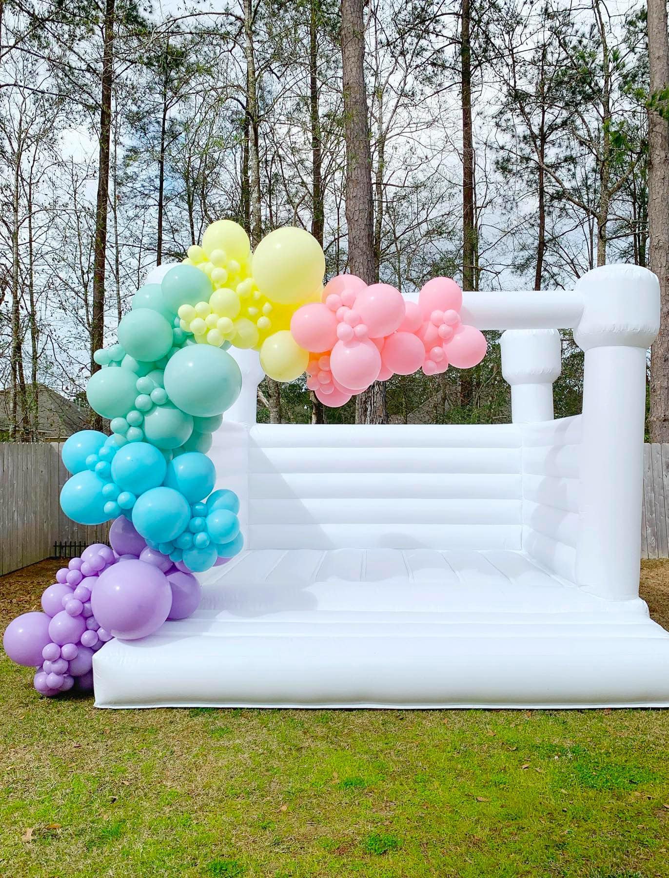 A large white inflatable bounce house is set up outside on a grassy yard for a rainbow baby shower. It is decorated with an arch of multicolored balloons, ranging from purple and blue to green, yellow, and pink. Tall trees and a wooden fence are visible in the background.