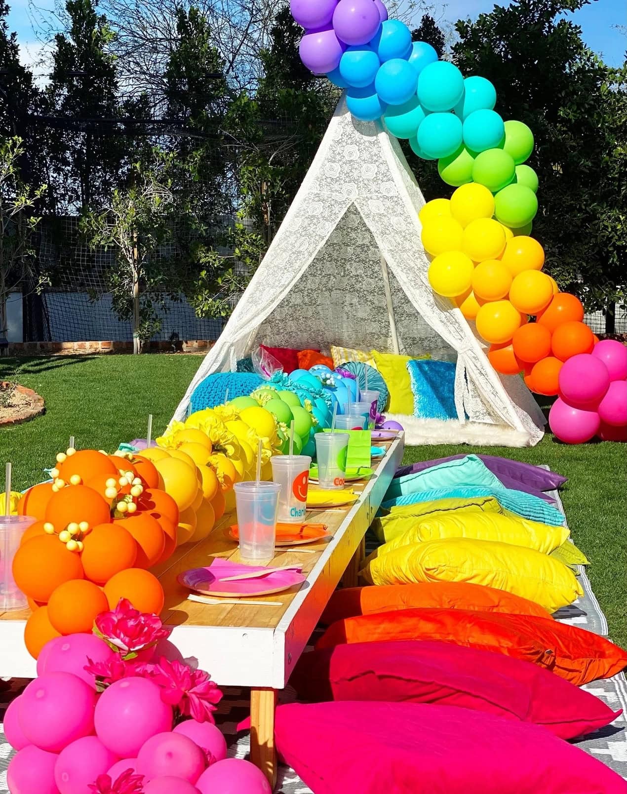 A festive outdoor picnic setup perfect for a rainbow baby shower features a low wooden table adorned with vibrant rainbow-colored balloons, pillows, and decorations. A white tent with lace fabric serves as the backdrop, with colorful cushions spread around. Drinks are placed on the table.