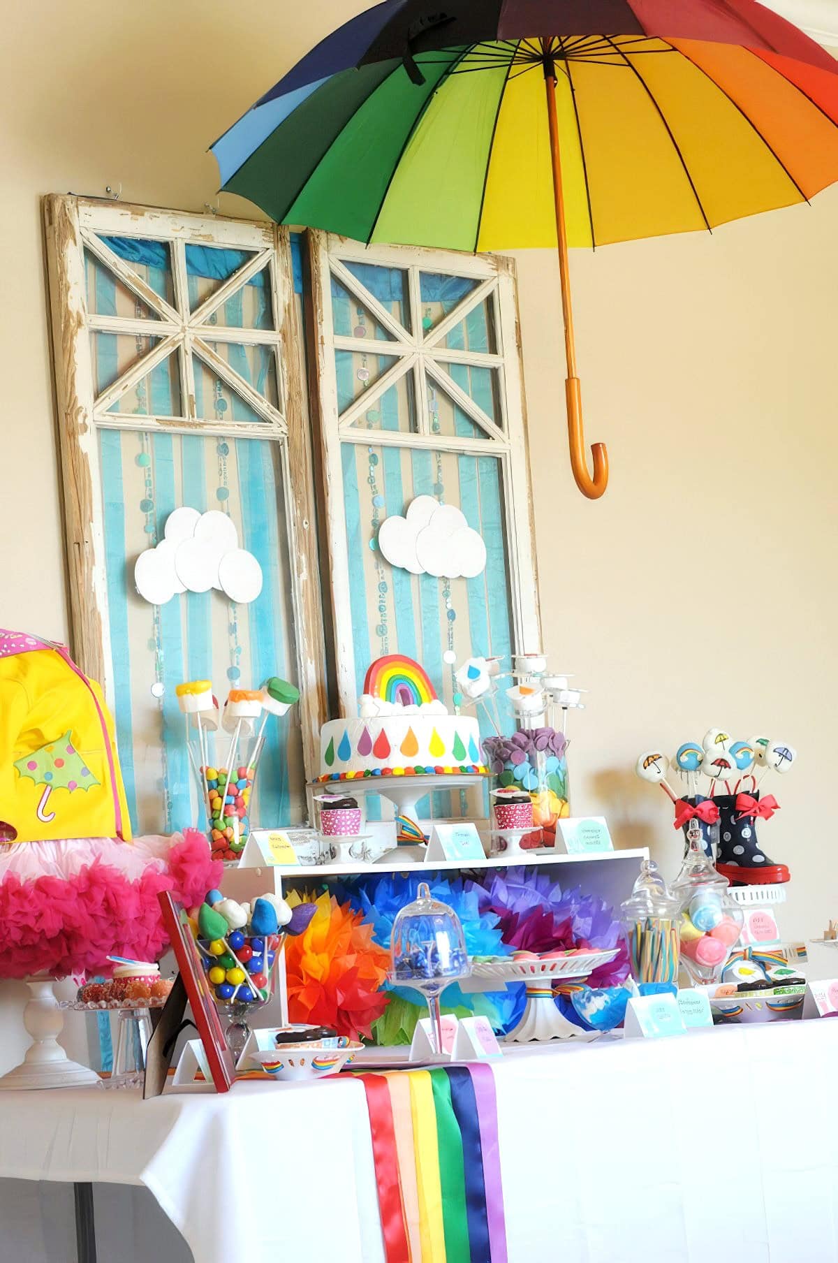 A colorful party setup with a rainbow theme perfect for a rainbow baby shower. Hanging from the ceiling is a rainbow umbrella, and the table below is decorated with rainbow-colored items, a multi-tiered cake with a rainbow design, various treats, and paper clouds. Rainbow banners adorn the table.
