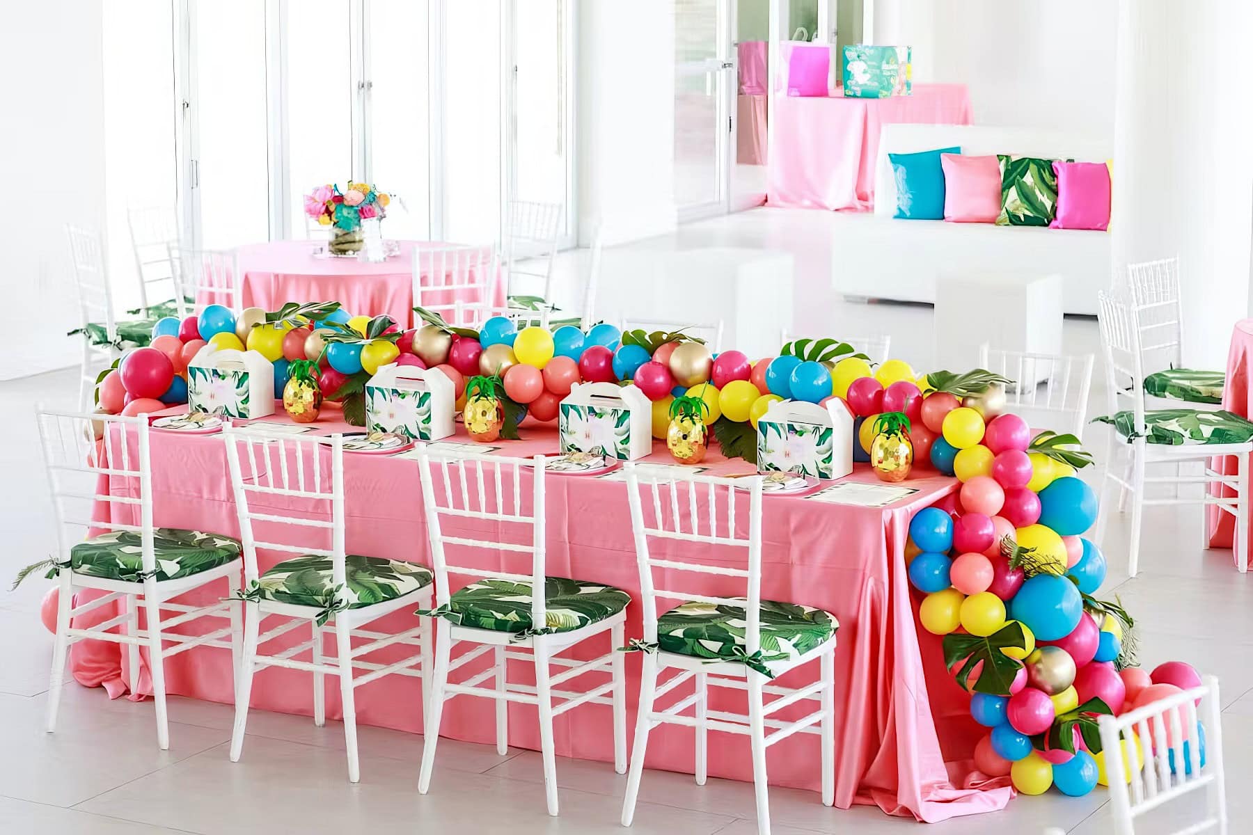 A vibrant party setup for a rainbow baby shower features a long table covered with pink cloth, white chairs with tropical leaf-patterned cushions, and a colorful balloon garland. The table is adorned with floral centerpieces and tropical-themed decorations. In the background are more party decorations.