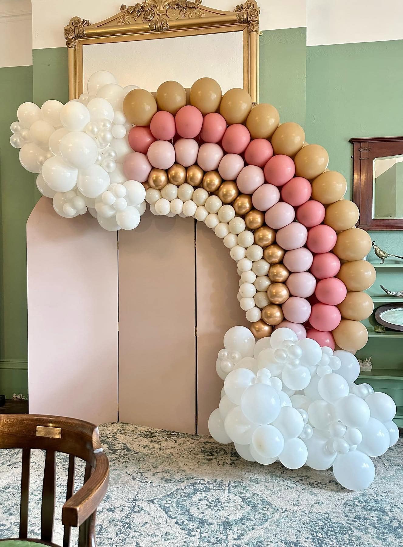 A colorful balloon arch is displayed against a pale pink wall, perfect for a rainbow baby shower. The arch consists of white, gold, pink, and peach balloons arranged in a rainbow shape. At the base, clusters of white balloons resemble clouds. A wooden chair is visible in the foreground.