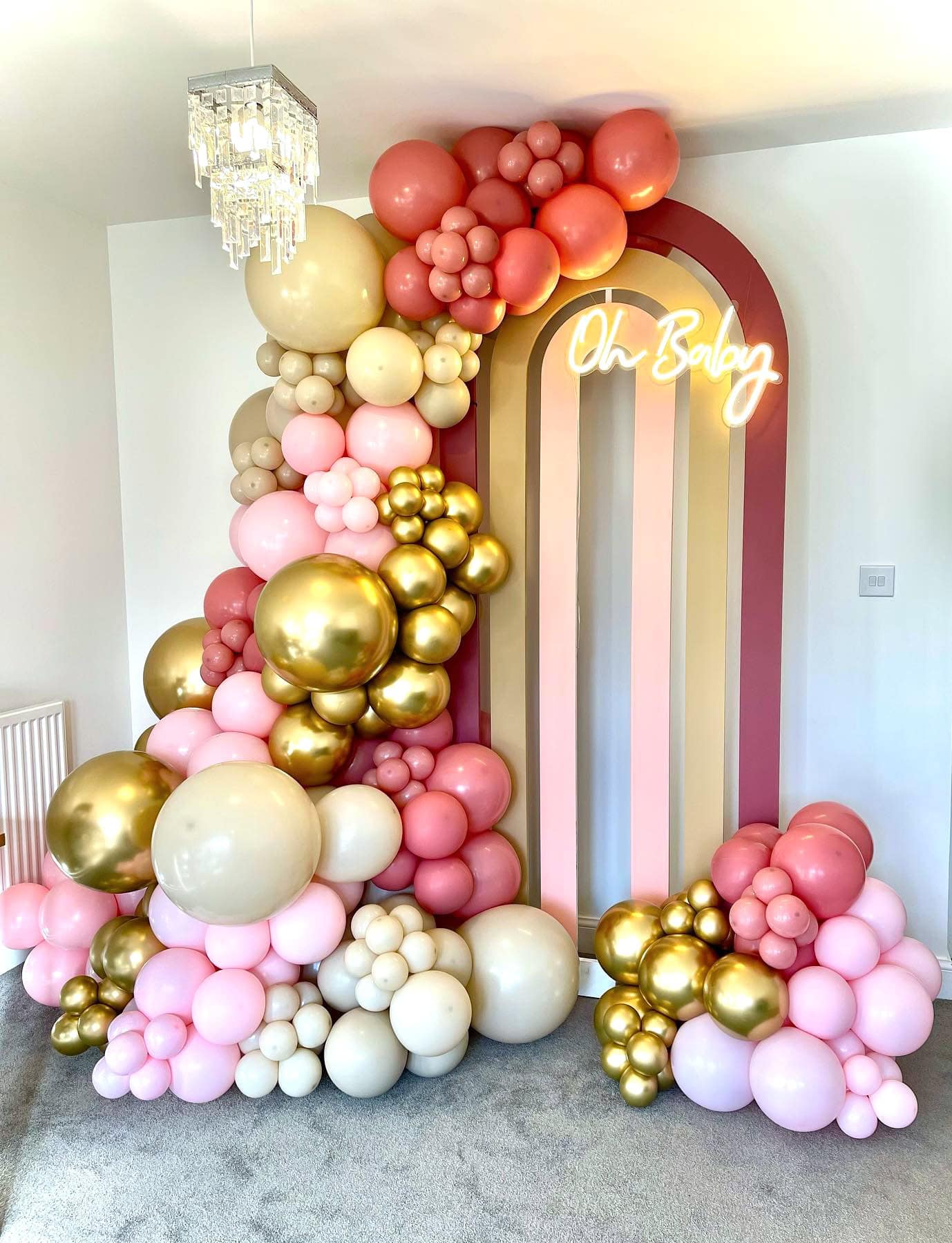 A colorful balloon arch featuring pink, gold, and white balloons adorns a corner of the room. The arch frames a striped backdrop and a neon sign that reads "Oh Baby," perfect for a rainbow baby shower. A chandelier hangs from the ceiling, and the room is painted white with gray carpet flooring.