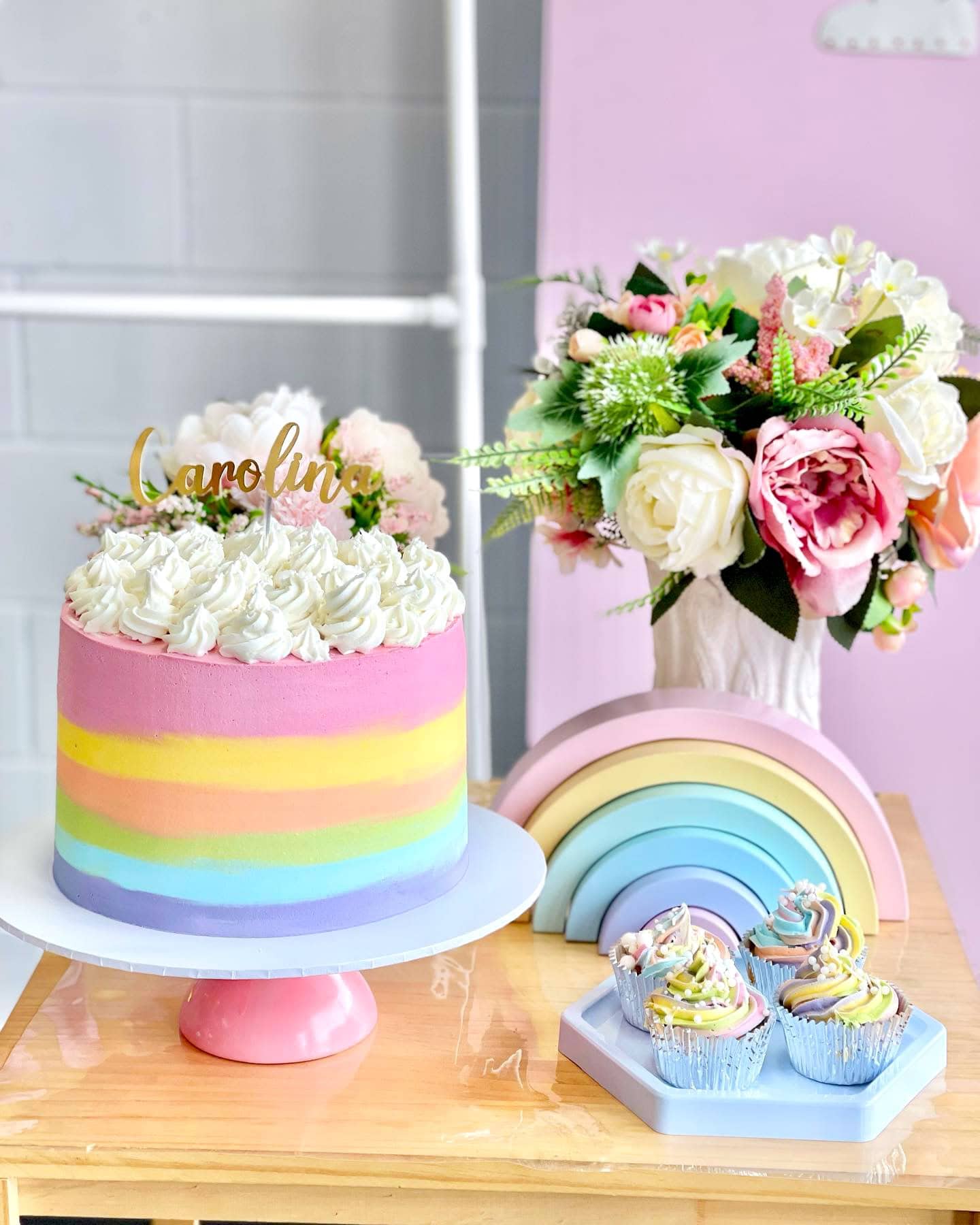 A pastel-colored rainbow-themed cake with a "Caroline" topper sits on a pedestal, perfect for a rainbow baby shower. Nearby are cupcakes with pastel swirls, a rainbow-shaped decoration, and a bouquet of pink and white flowers. The background features a light pink and grey wall.