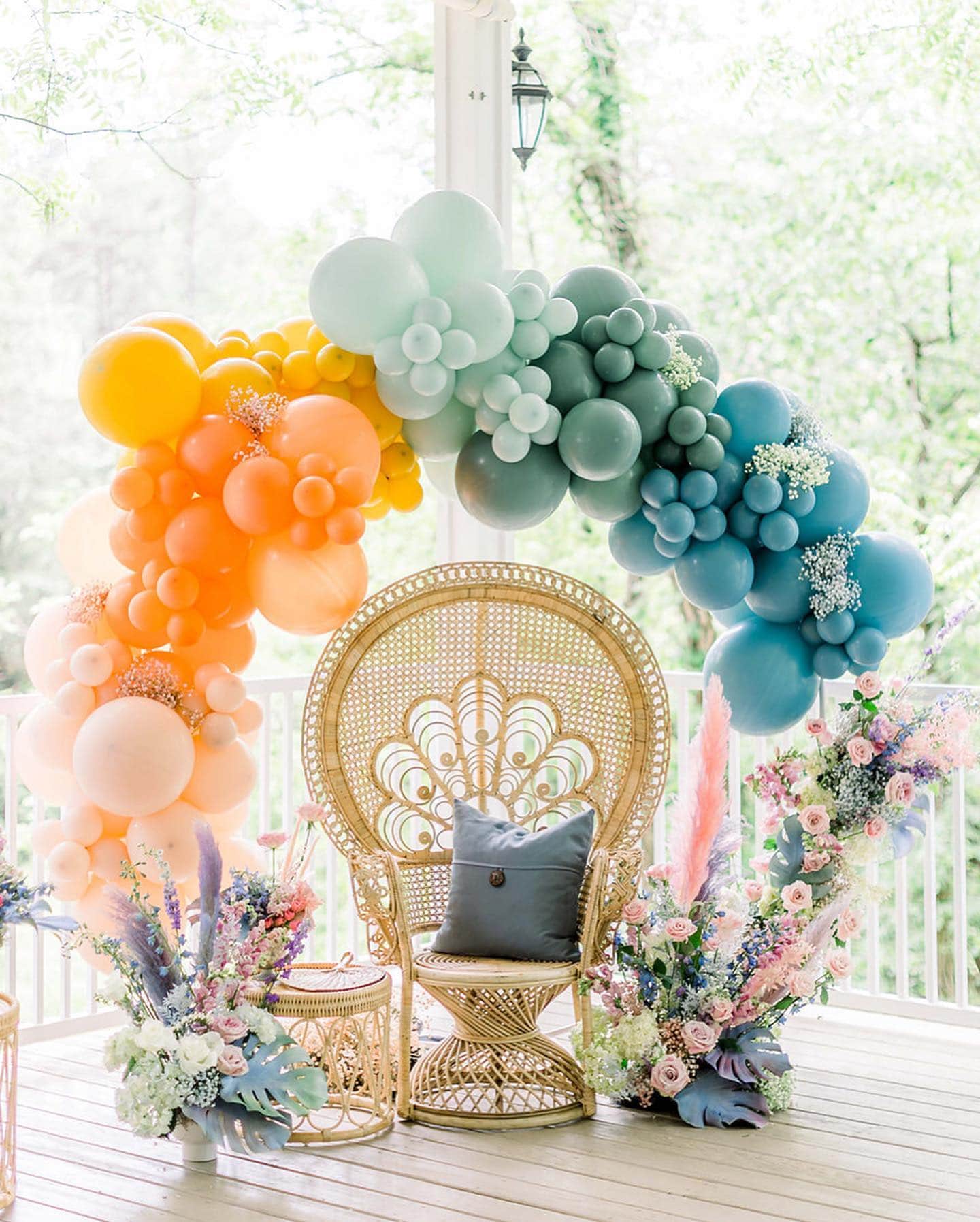 A wicker chair with a dark cushion is centered under a balloon arch in pastel shades of orange, green, and blue. Vibrant floral arrangements surround the setup on both sides, creating a festive and elegant scene perfect for a rainbow baby shower.