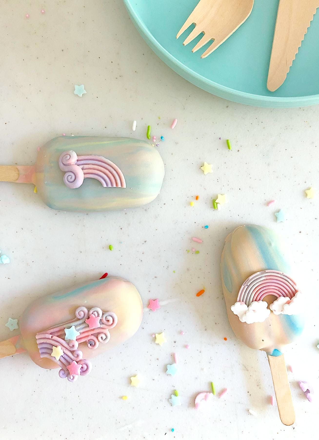 Three pastel-colored decorated cakesicles with rainbow and cloud decorations are placed on a white surface, perfect for a rainbow baby shower. Pastel star and sprinkle decorations are scattered around. A turquoise plate with wooden utensils is in the top right corner.